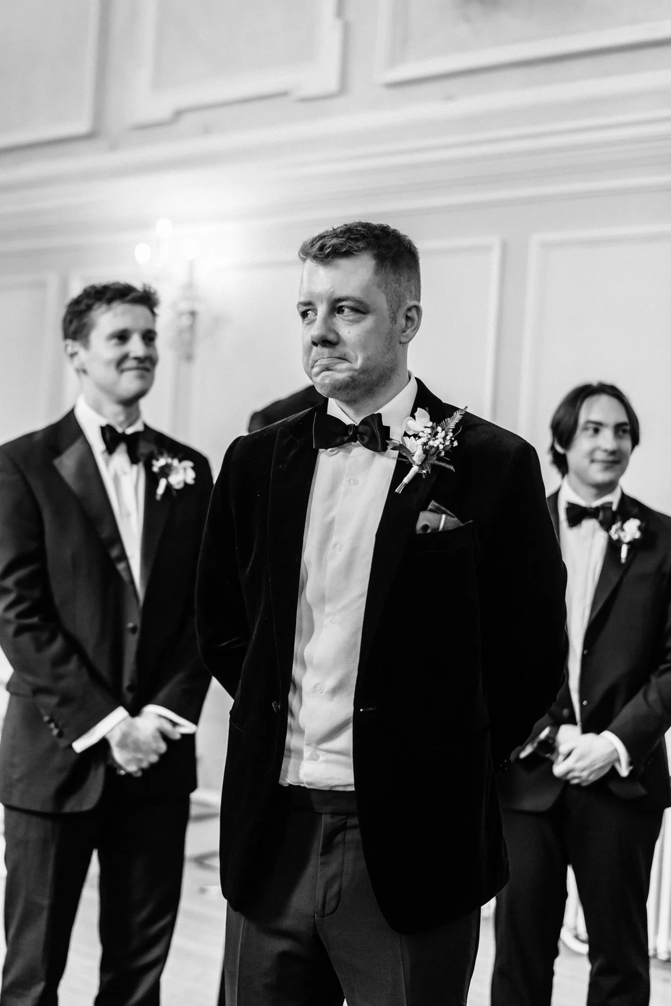 Black and white photo of a groom with a boutonniere, displaying an emotional expression during a wedding ceremony, standing among groomsmen dressed in tuxedos and bow ties.