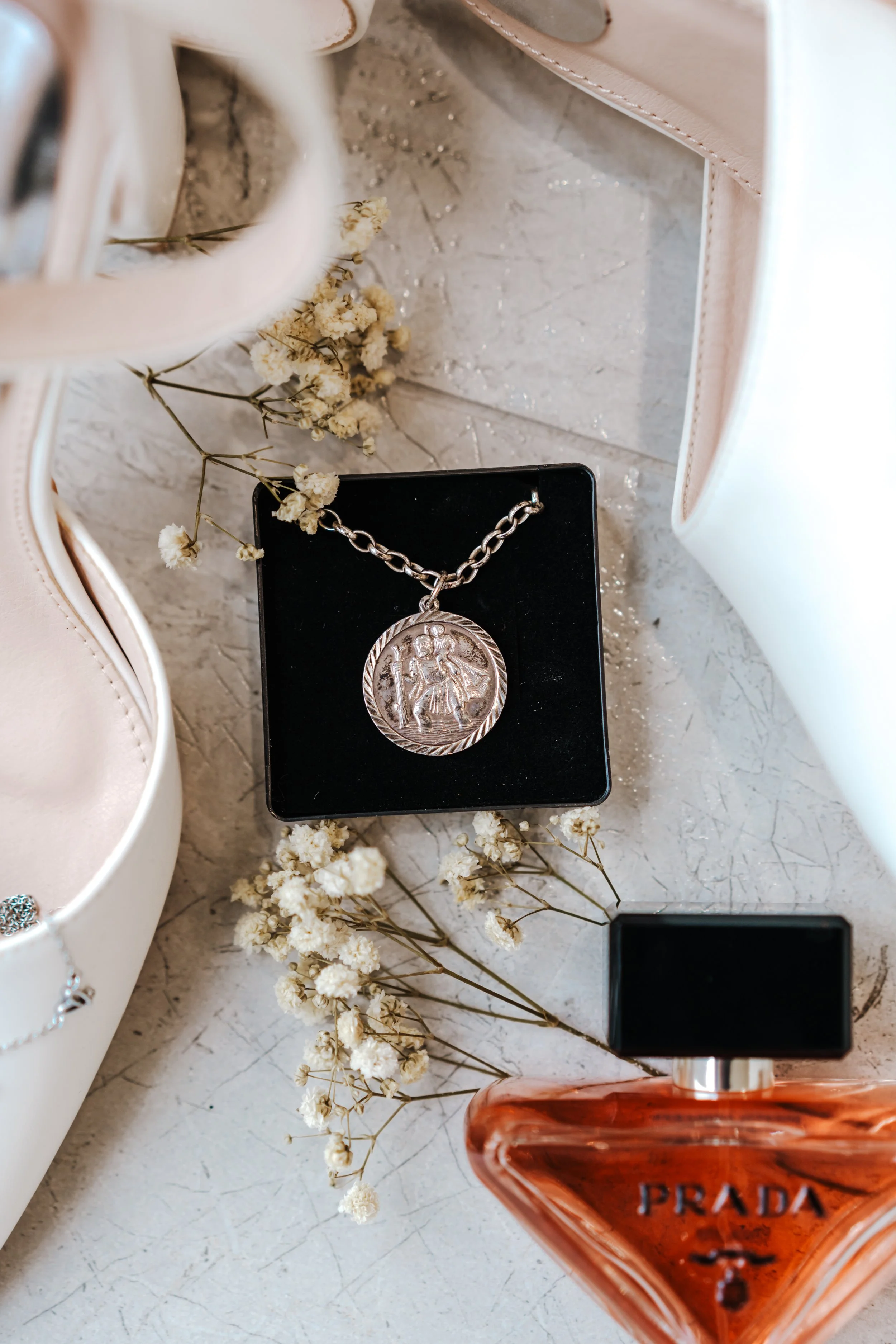 A silver pendant necklace with a religious figure design on a black jewelry box, surrounded by dried white flowers, near a pair of light-colored high heels, and a bottle of Prada perfume on a light surface.