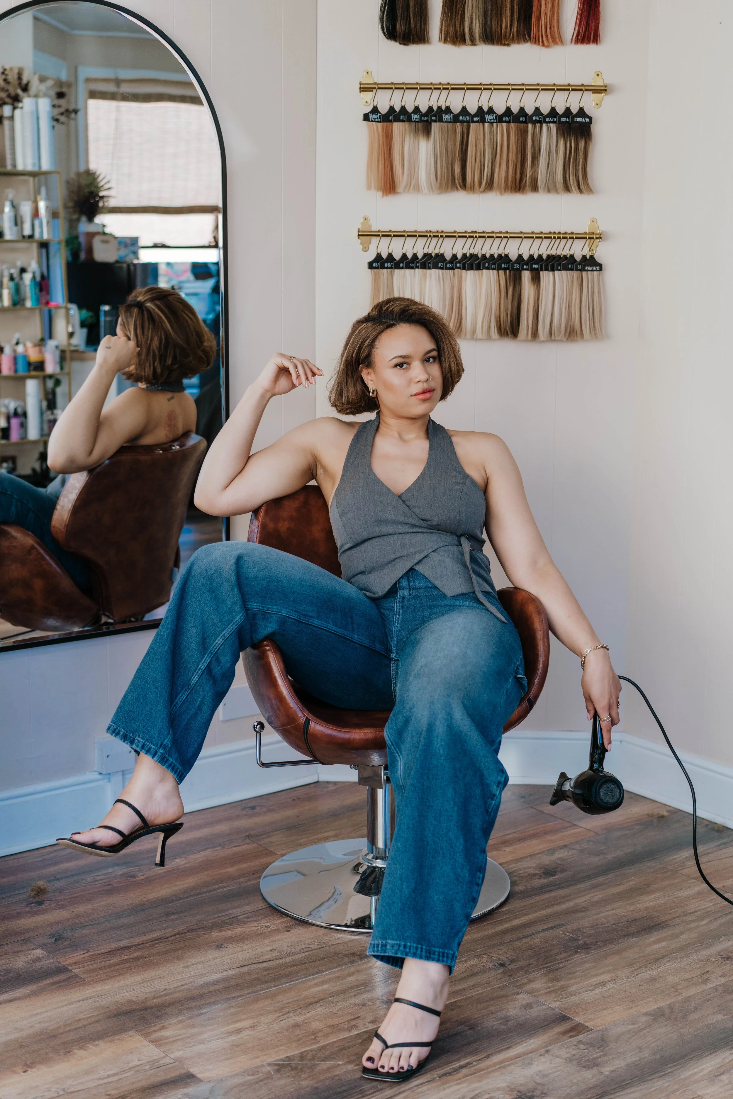 A woman sitting in a salon chair, flexing her right arm and looking at the camera. She has shoulder-length brown hair and is wearing a gray sleeveless top, blue jeans, and black strappy high heels. Behind her are hair color samples hanging on wall-mounted racks, and a large mirror reflected a shelf with hair products and a window with blinds.