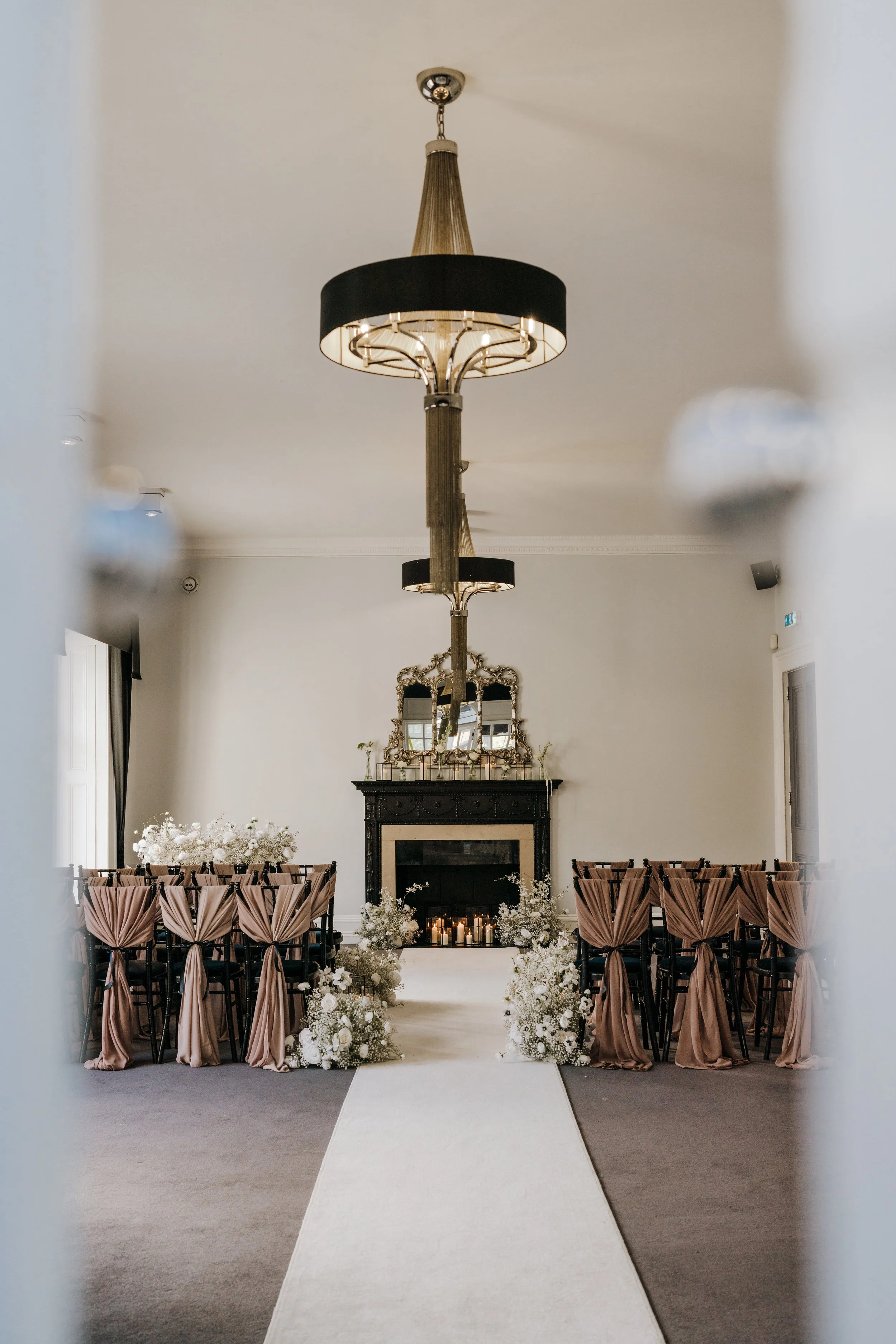 Elegant wedding ceremony setup with a white aisle runner, floral arrangements, and chairs draped in pink fabric, leading to a fireplace with candles and a decorative mirror above it.