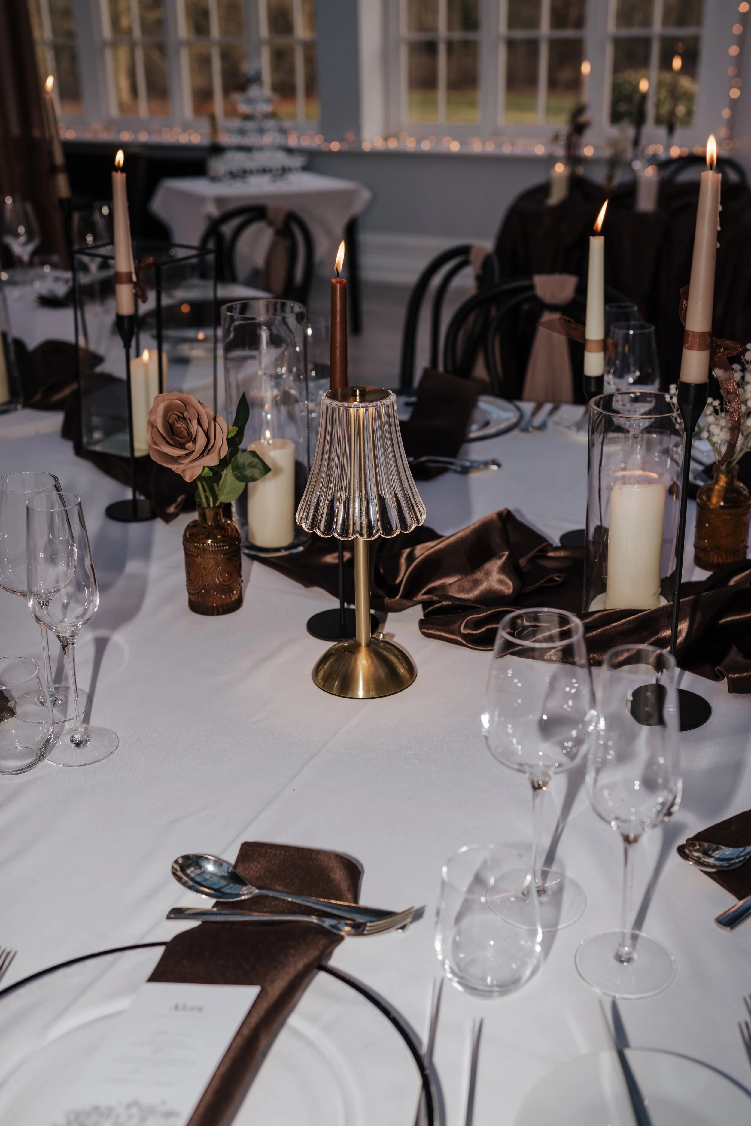 Elegant dining table decorated with candles in glass holders, a pink rose in a small gold vase, a brass lamp with a pleated shade, and dark napkins, set for a formal event.