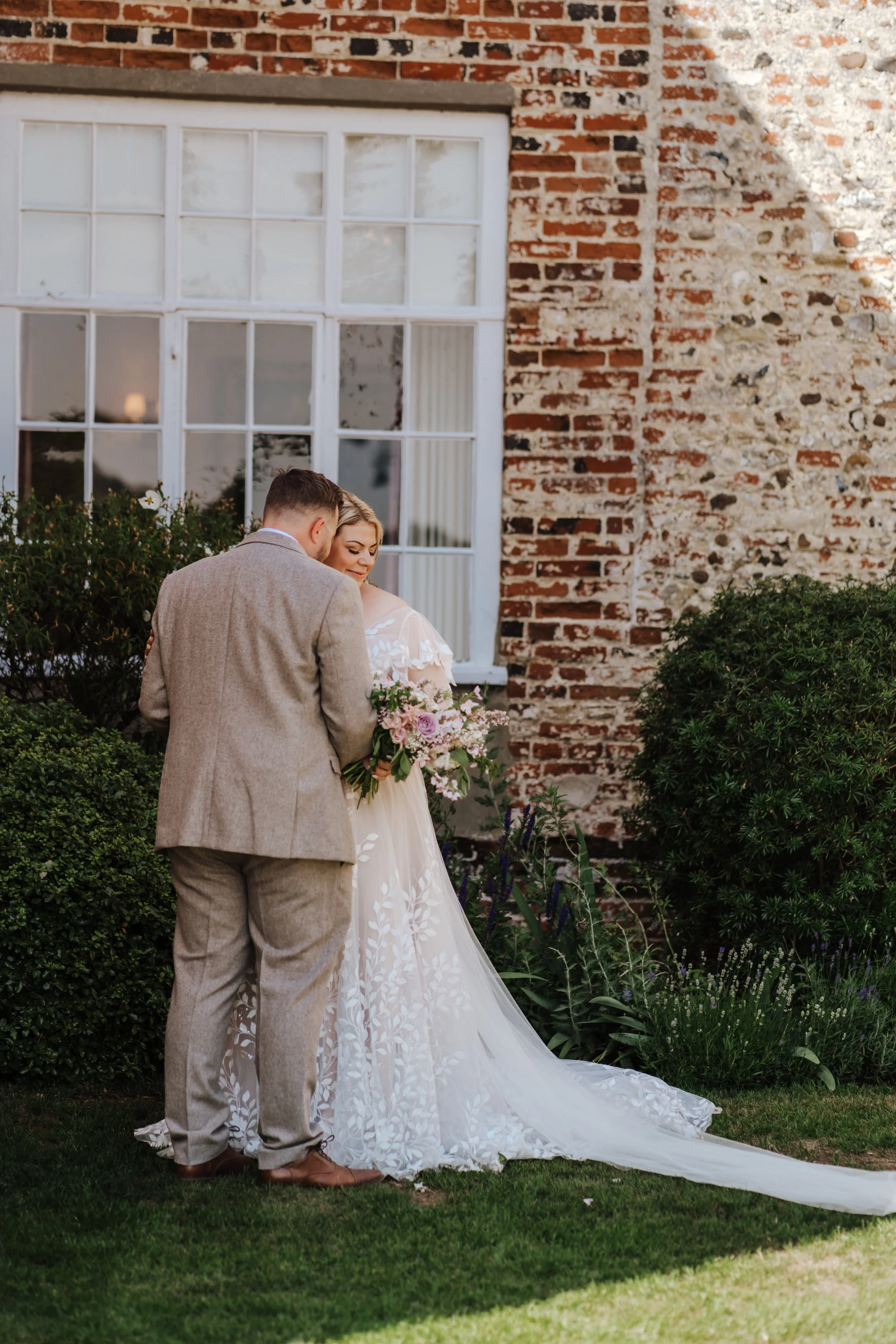 A bride and groom standing close together outside in front of a brick building and large window, with the bride holding a bouquet of pink and purple flowers.