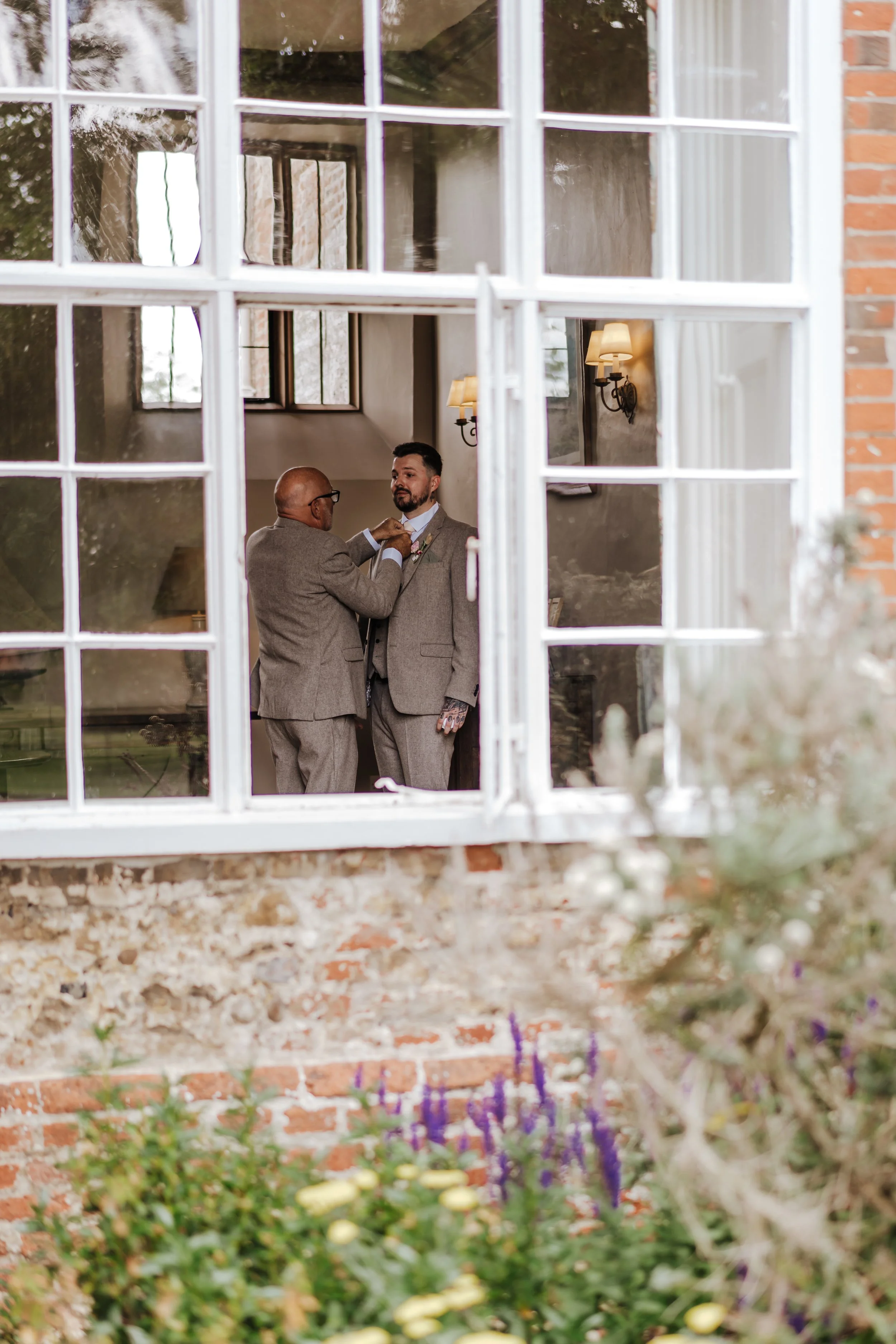 A man helps another man adjust his suit jacket inside a room, viewed through a window with multiple panes, with soft indoor lighting and exterior flowers visible.
