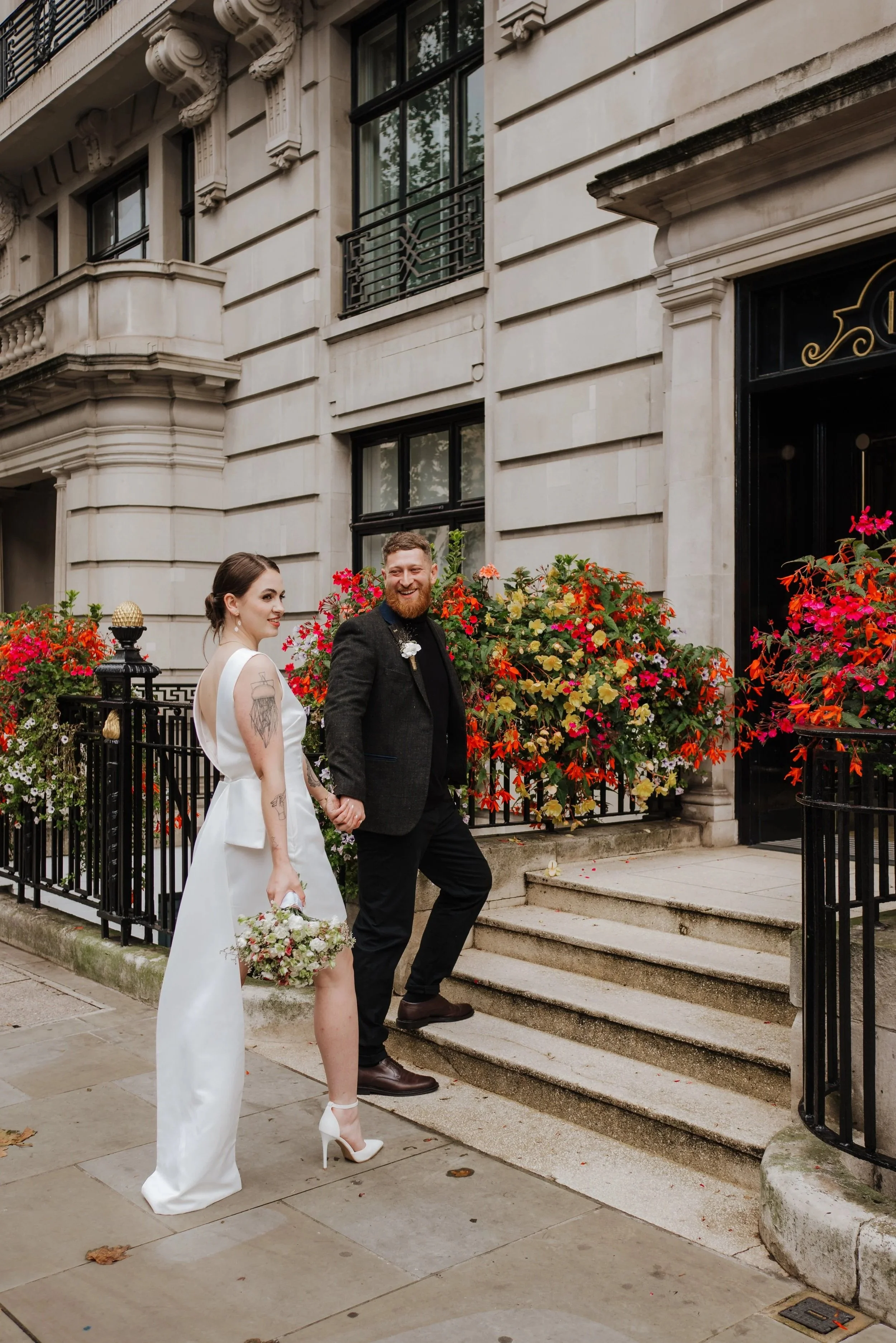 A joyful couple standing outside a historic building, holding hands, with the woman holding a bouquet, surrounded by flower arrangements on the porch.