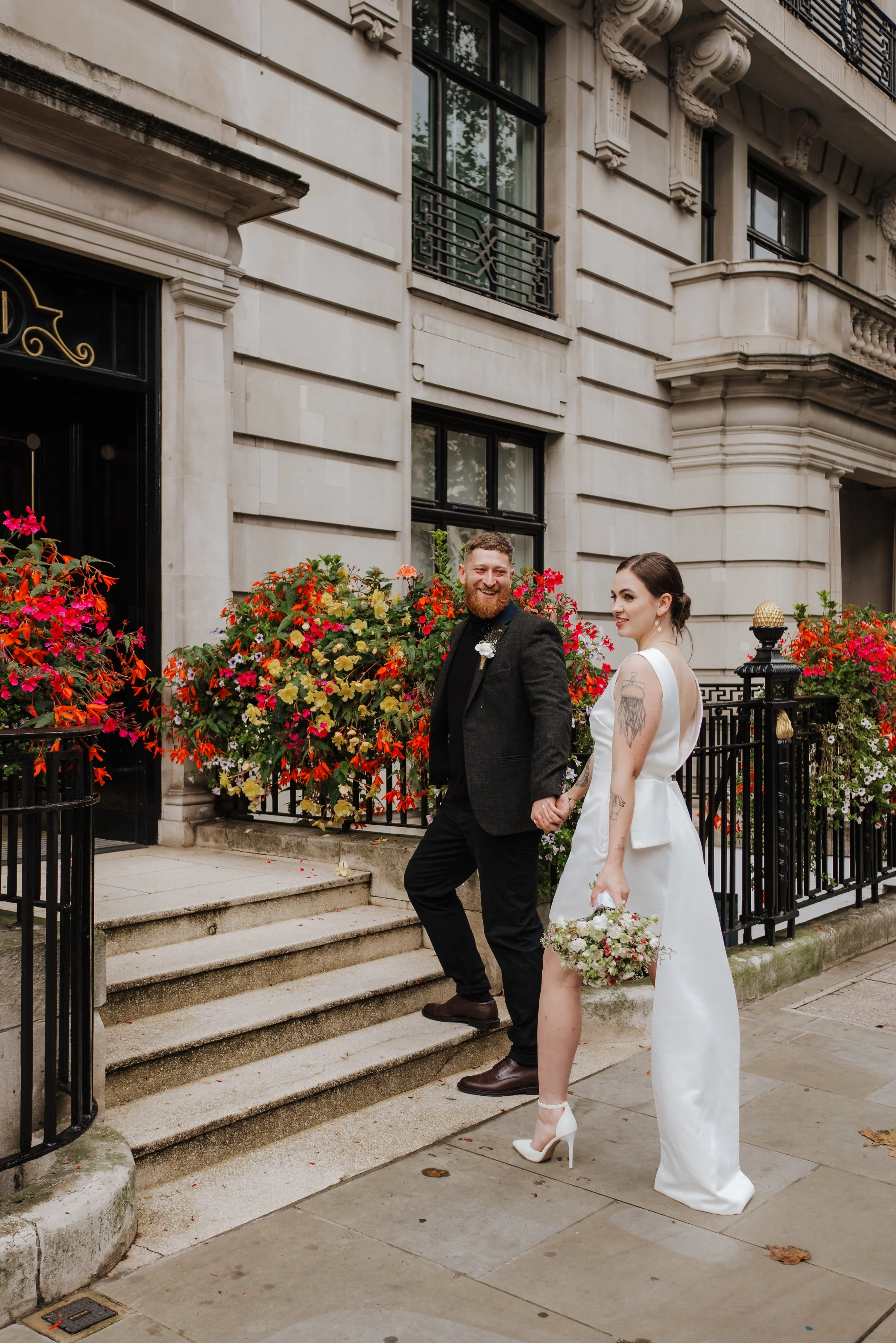 A happy couple in wedding attire holding hands outside a historic building with colorful flower arrangements.