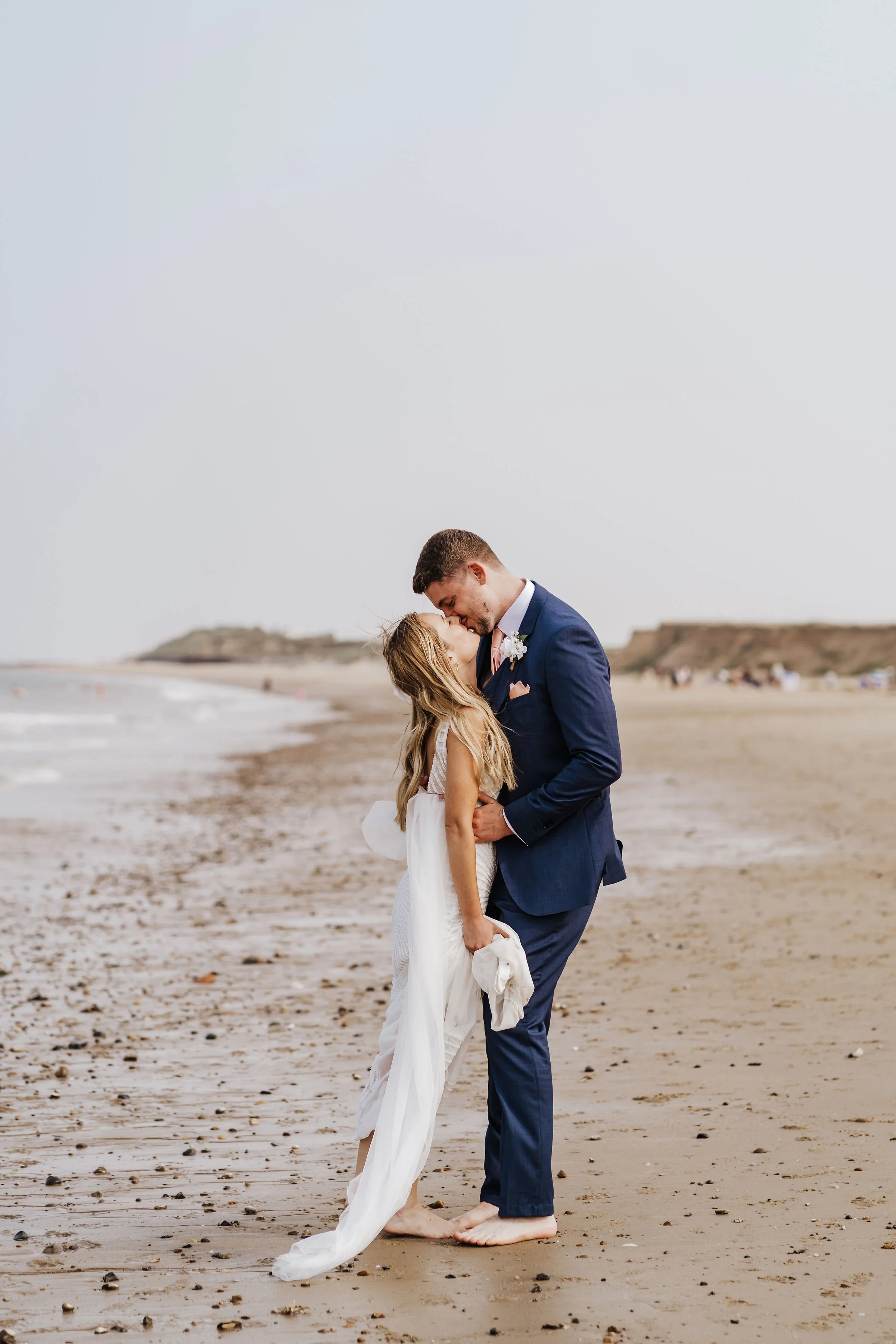 A couple in wedding attire sharing a kiss on the beach with sandy shores, ocean waves, and distant cliffs in the background.