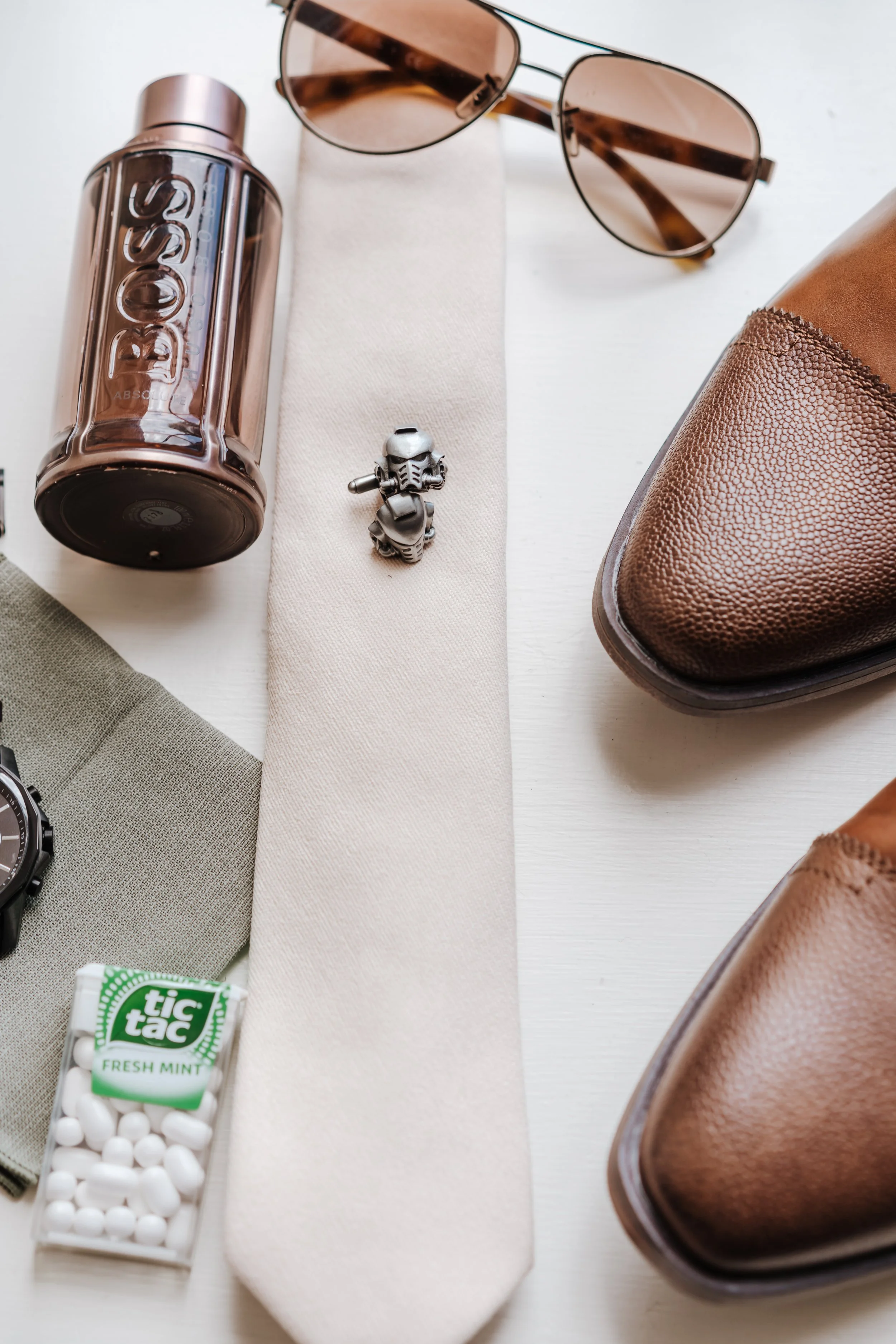 Flat lay of men's accessories and shoes on a white surface, including sunglasses, a metal robot figure, a bottle of Boss cologne, a watch, gum, and brown dress shoes.