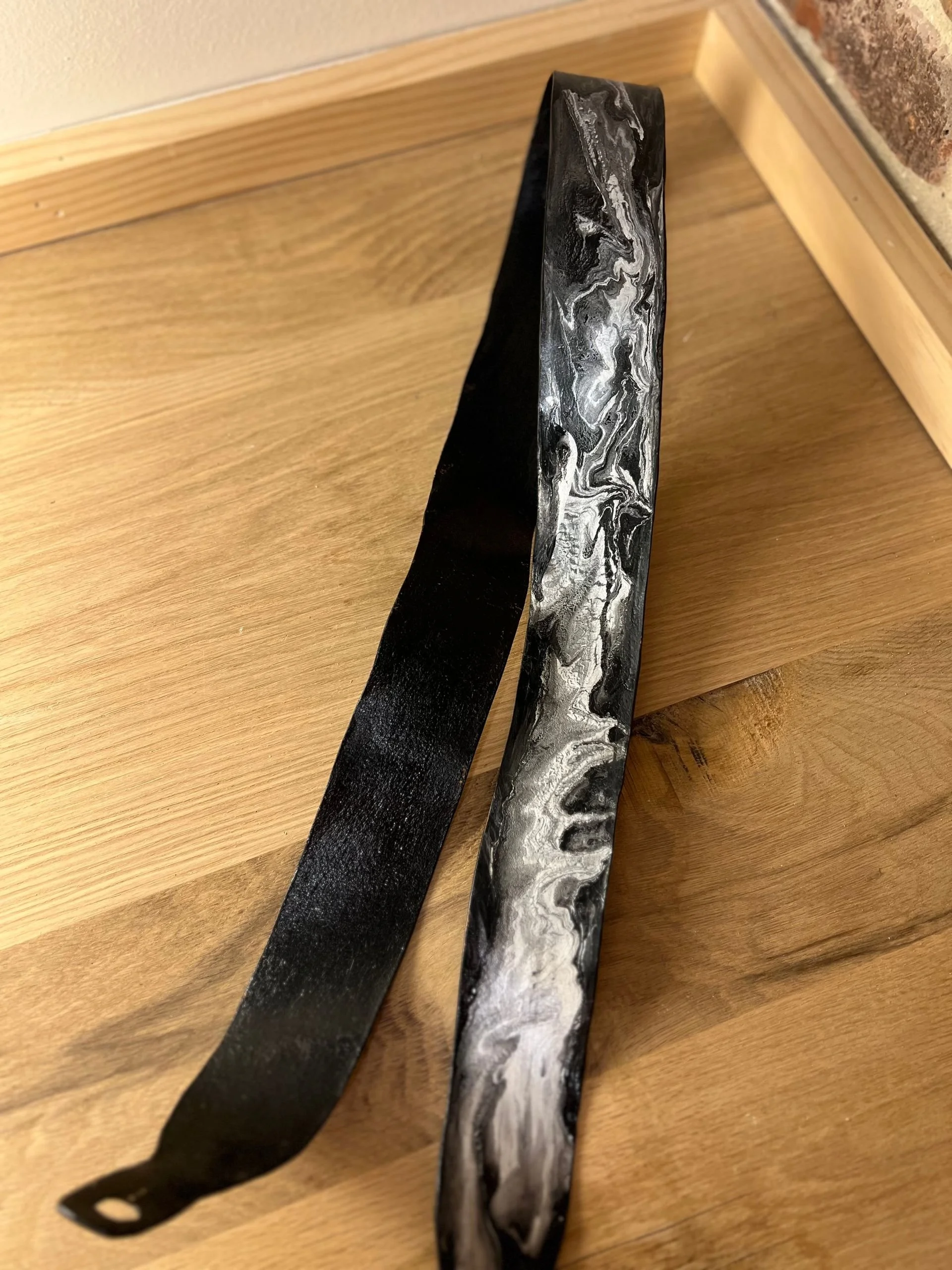 A black and white marbled leather belt placed on a wooden surface near a light-colored wall and a wooden baseboard.