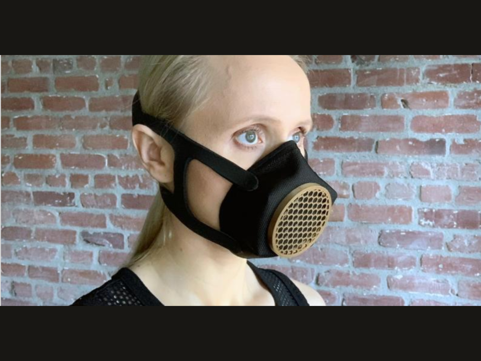 A woman with blonde hair wearing a black face mask with a filter cartridge in front of a brick wall.