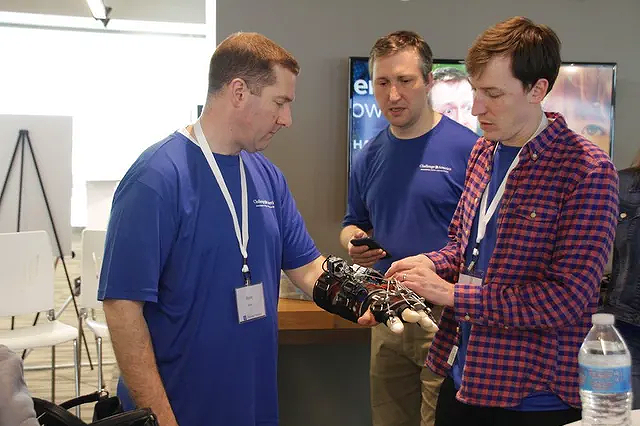 Three men, two in blue shirts and one in a red checkered shirt, are examining a prosthetic limb with a robotic hand. They are indoors at a conference or event with white chairs and a large screen in the background.