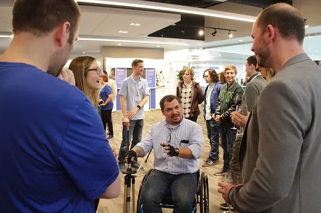 A man in a wheelchair demonstrates a device to a group of people in an indoor setting.
