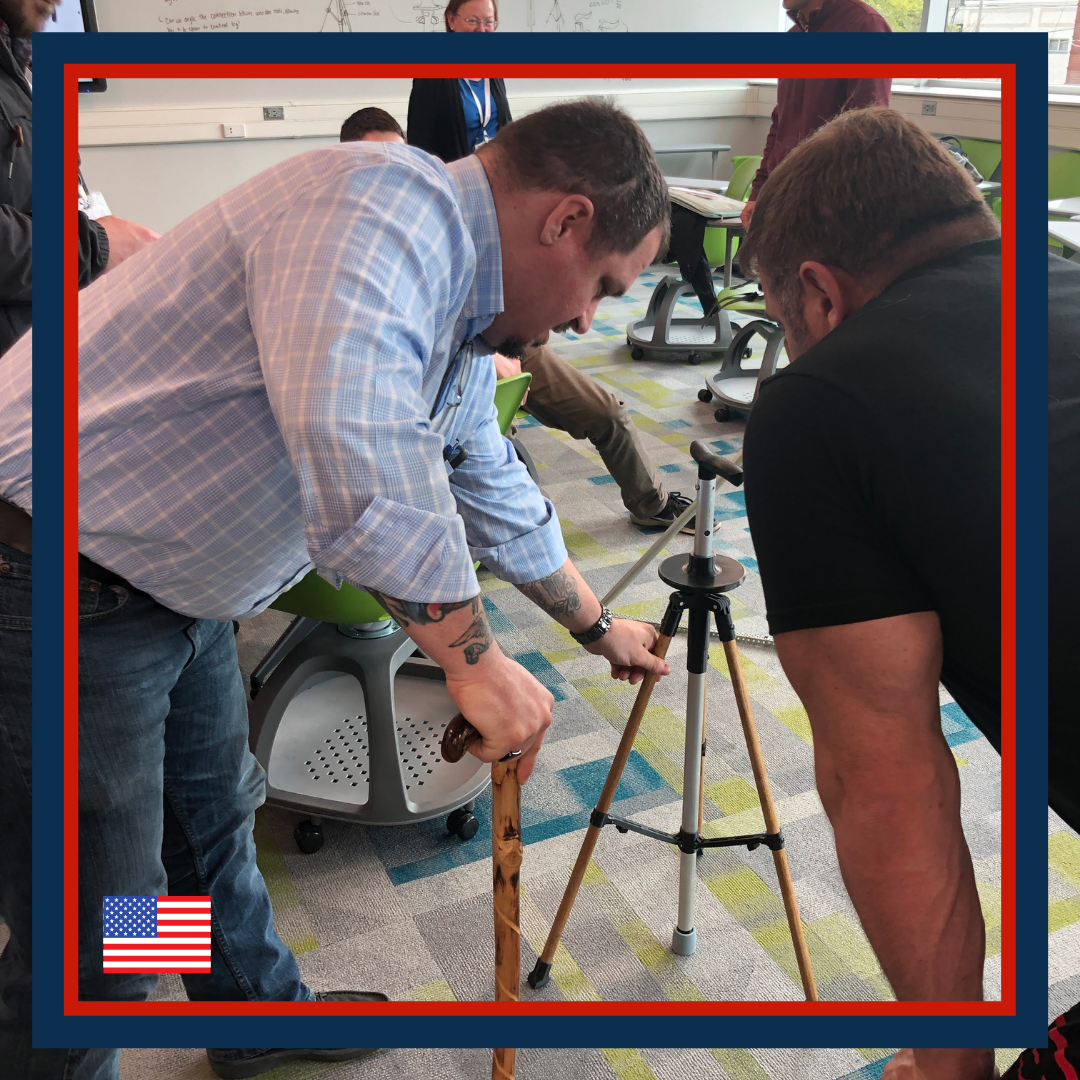 Two men working together to assemble a tripod or surveying equipment in a classroom with colorful carpet and green chairs.