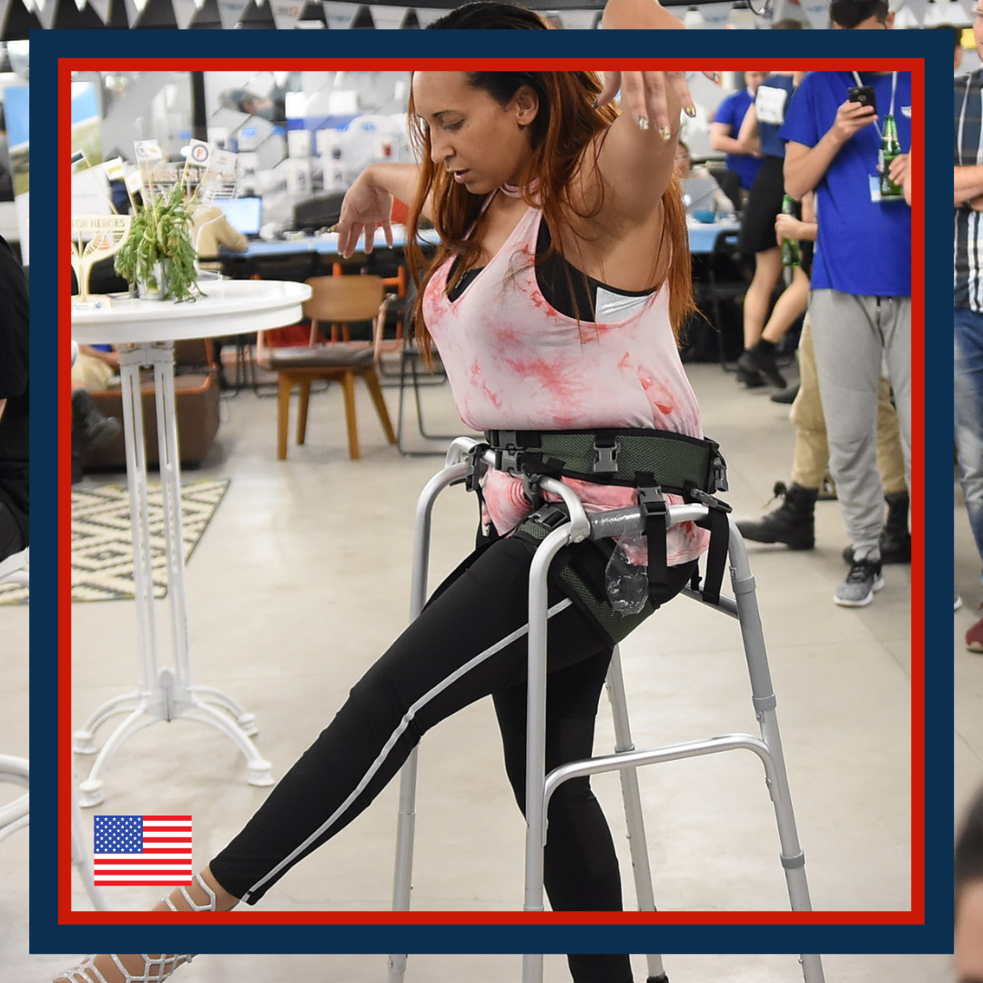 A woman in a pink tie-dye tank top and black pants using a walker at an indoor event with several people in the background.