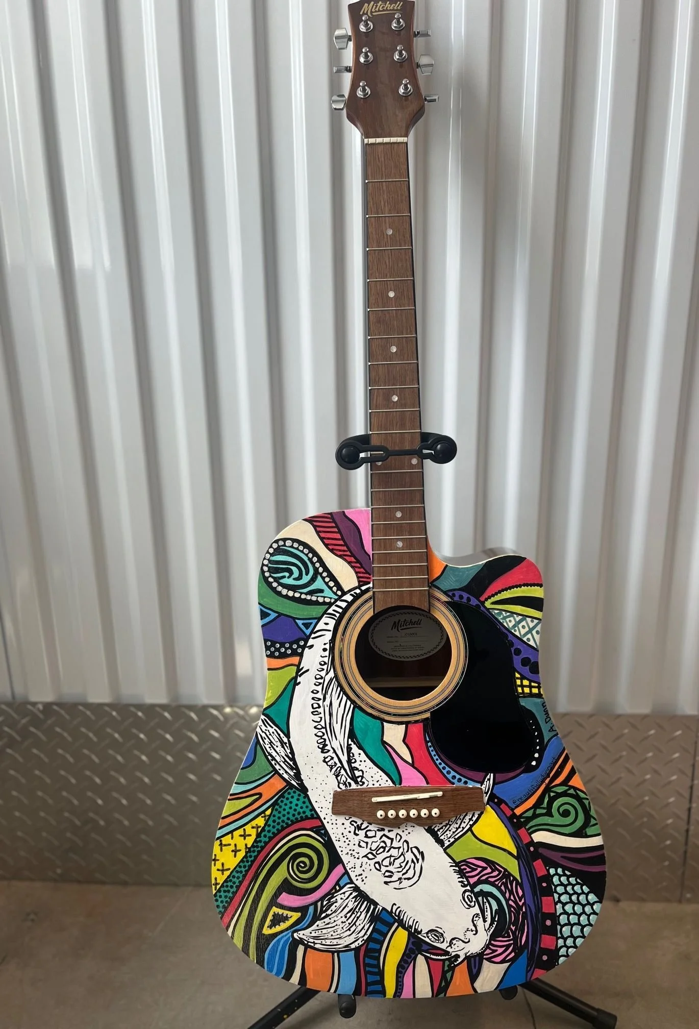 An acoustic guitar with colorful, abstract artwork featuring a stylized animal, possibly a rabbit, painted on the body. The guitar has a wooden neck and headstock, and it is standing upright on a black stand against a metallic, ribbed wall background