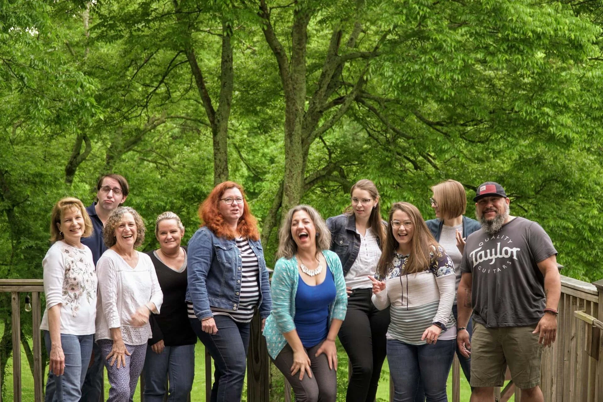 Group of ten people standing on a wooden deck outdoors with green trees in the background, smiling and laughing together.