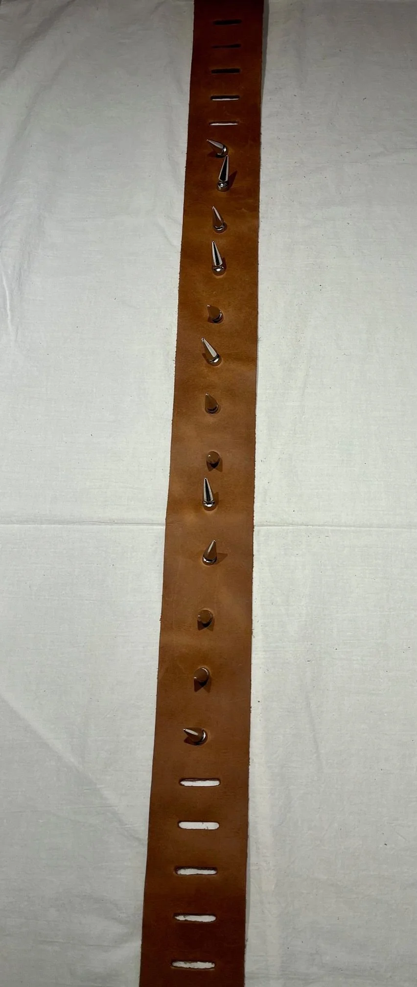 A vertical strip of brown leather with various types of metal tacks or studs pressed into it, laid flat on a white surface.
