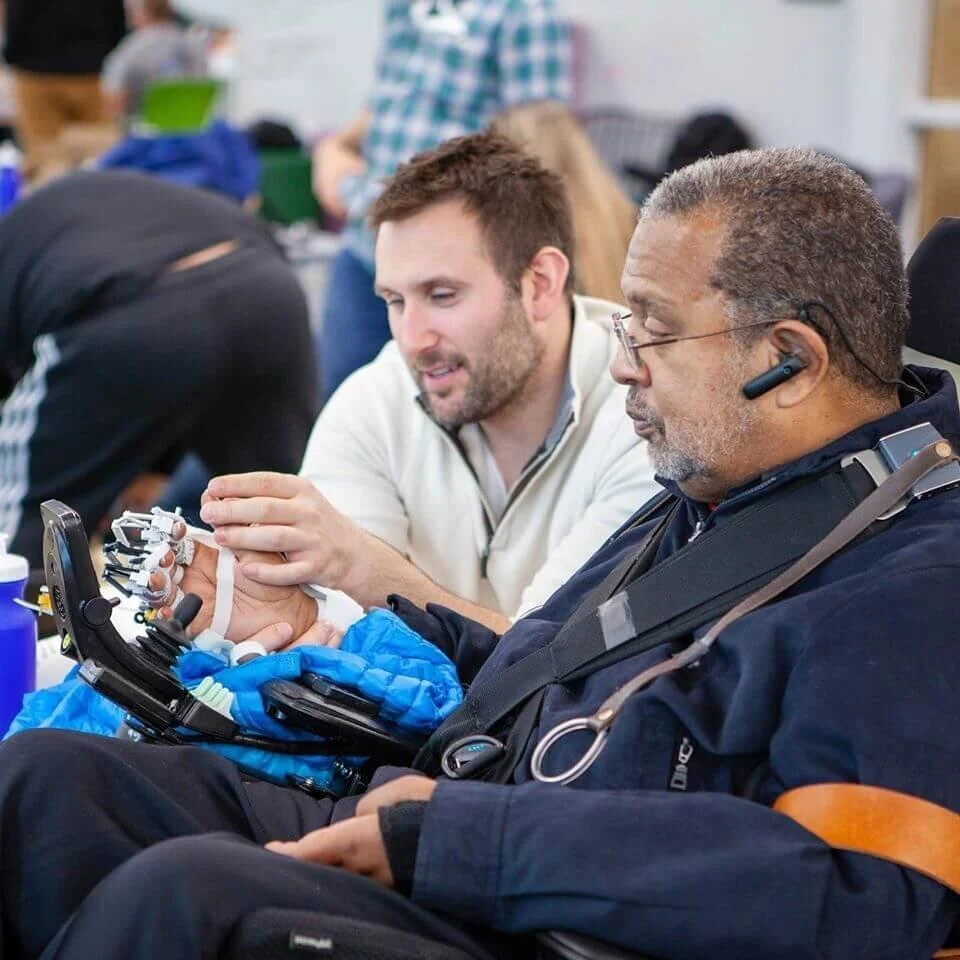 A young man assists an older man with a prosthetic hand at a public event or workshop, both focused on the prosthetic and its features.
