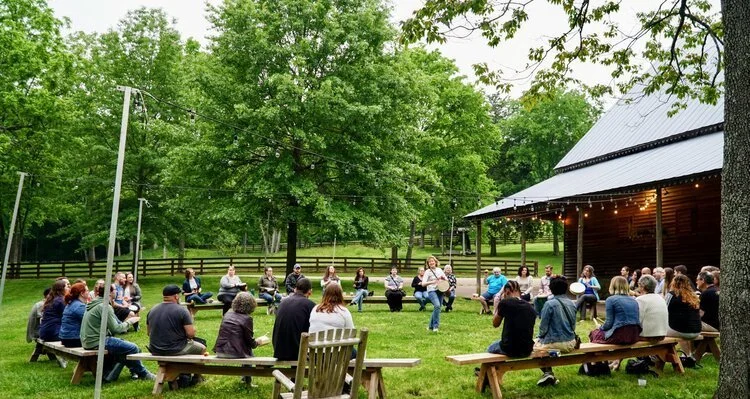 People sitting in a circle outdoors on wooden benches during a gathering or event, with a large tree, a building with string lights, and lush green trees in the background.
