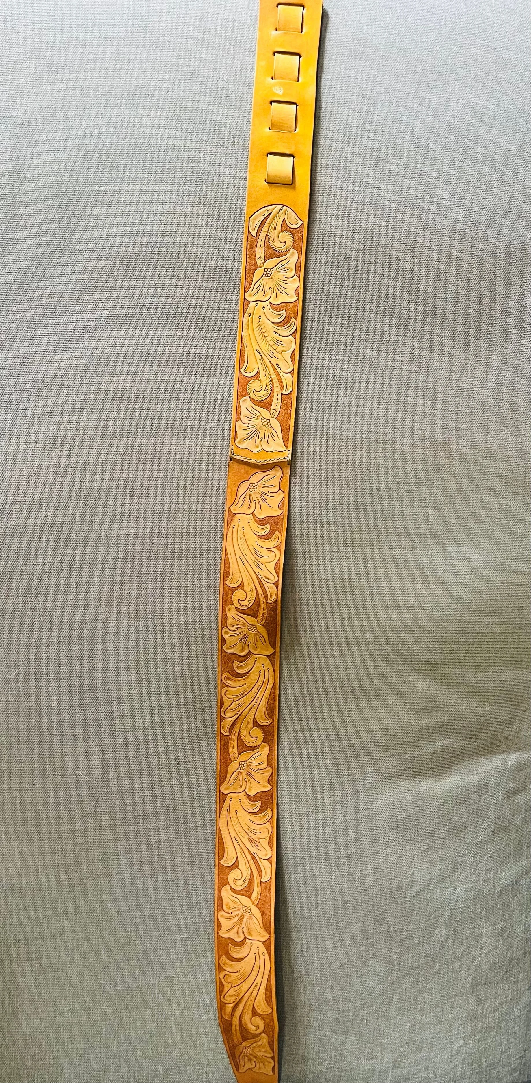 A leather belt with floral carved designs, hanging against a gray fabric background.