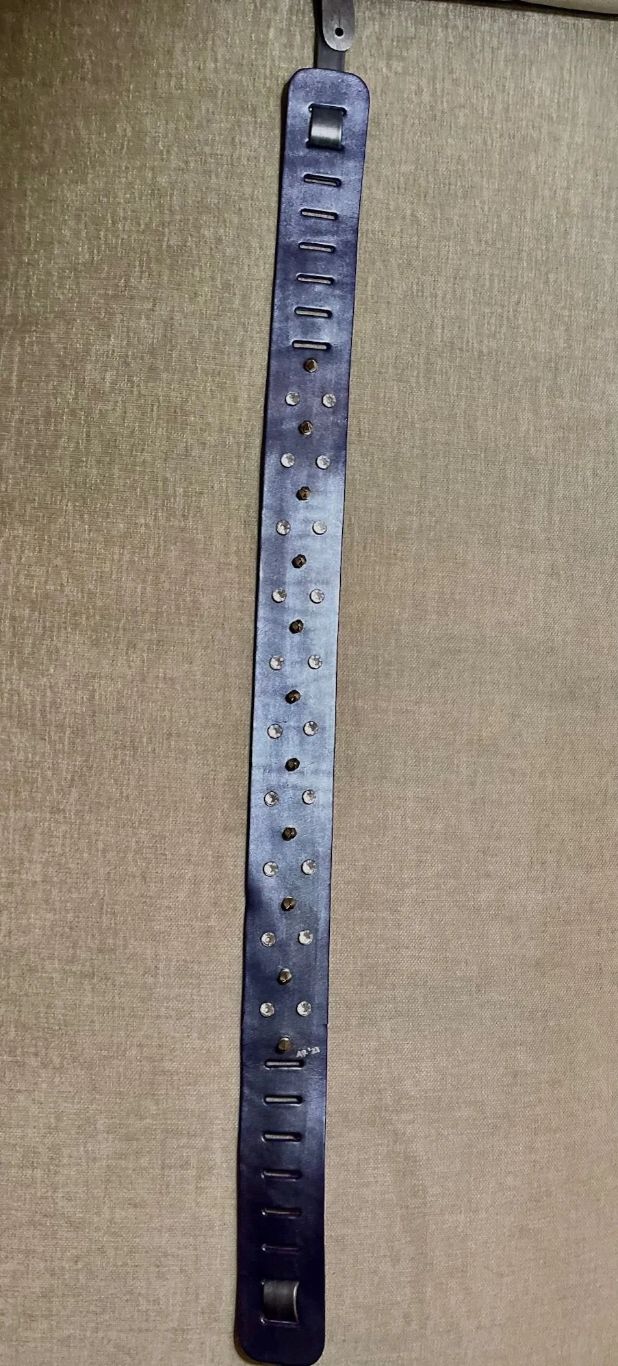 A medical metal gait belt with multiple holes, adjustable buckle, and slots for grip, hanging vertically against a beige background.