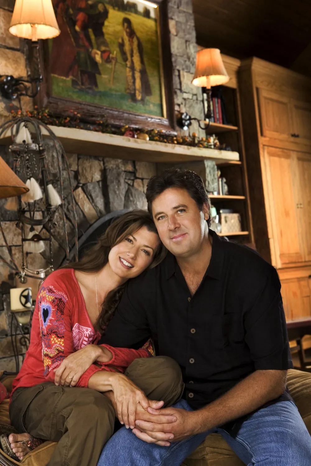 A man and woman sit close together inside a cozy, rustic living room with a stone fireplace, wooden shelves, and warm lighting. The woman wears a red patterned top, and the man wears a black shirt.