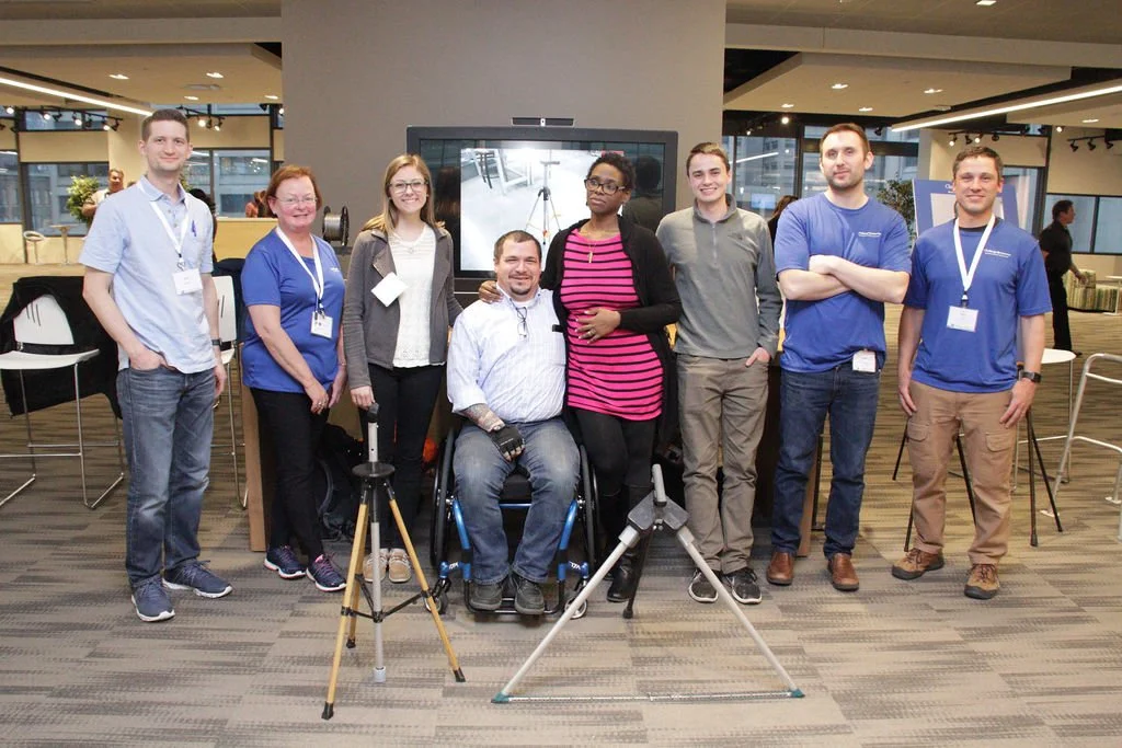Group of eight people at a professional event, some wearing name badges, standing in an office or conference space. There are cameras and equipment in front of them, with a display screen behind.