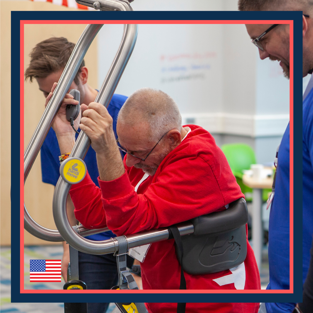A man in a red sweatshirt participating in a rehabilitation or therapy session assisted by two therapists, one in a blue shirt, in an indoor facility. The man is using a gait training device.