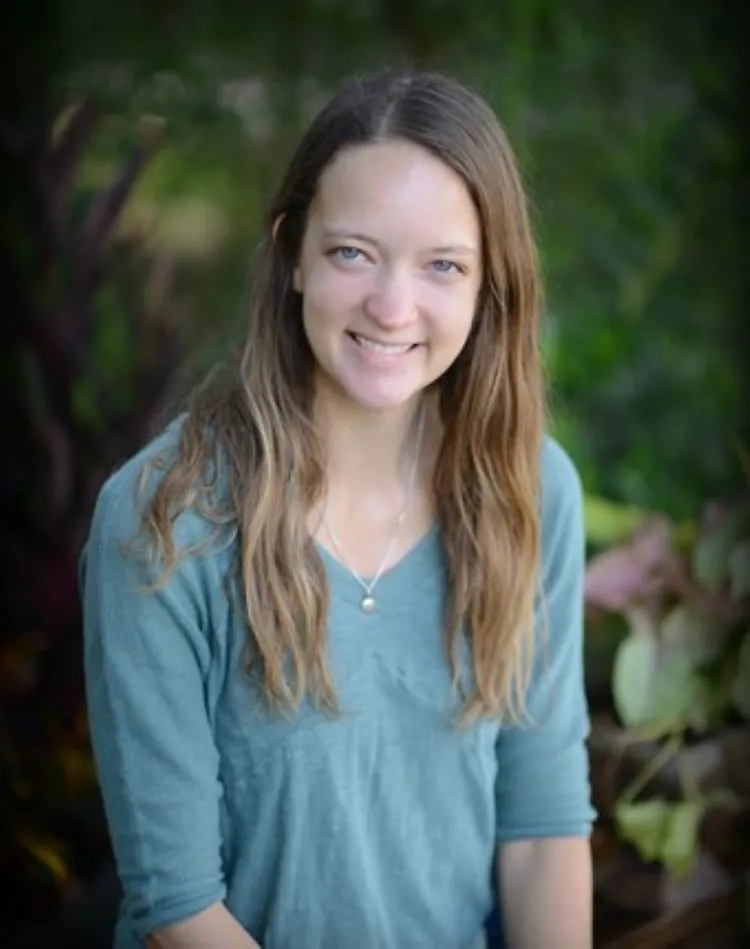 A young woman with long wavy brown hair and blue eyes, smiling, wearing a light blue shirt and a necklace with a pearl, outdoors with green foliage in the background.