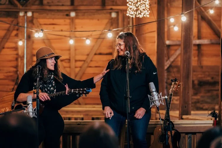 Two women with guitars performing in a rustic wooden barn decorated with string lights, one singing into a microphone and the other gesturing with her hand.