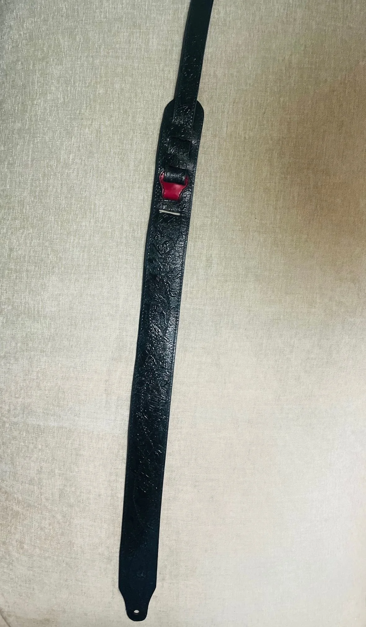 A black leather dog leash with a red metal clip, hanging vertically against a beige textured background.