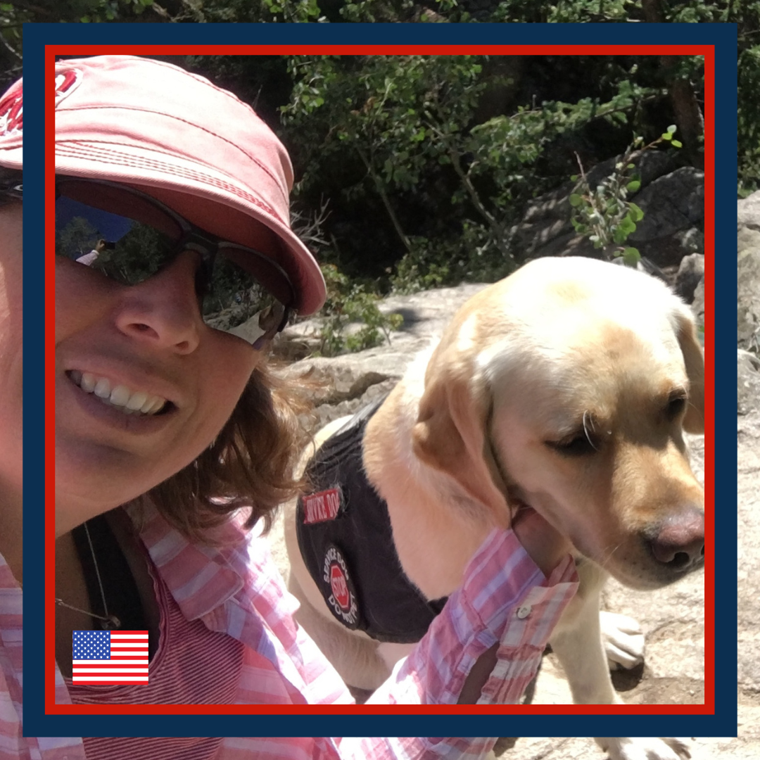 A woman in sunglasses and a pink hat smiling at the camera, holding a yellow Labrador retriever dog with a black vest on a rocky outdoor trail surrounded by green bushes.
