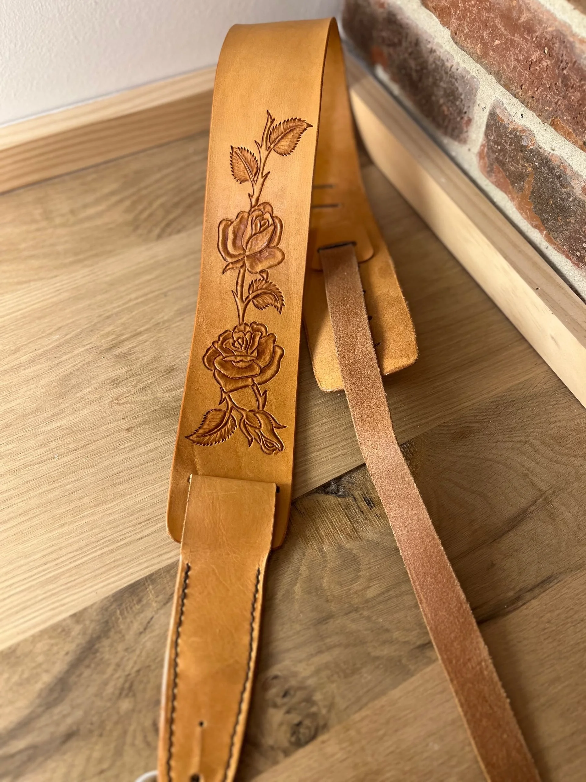 Tan leather belt with an embossed floral design of roses and leaves, placed on a wooden surface next to a brick wall.