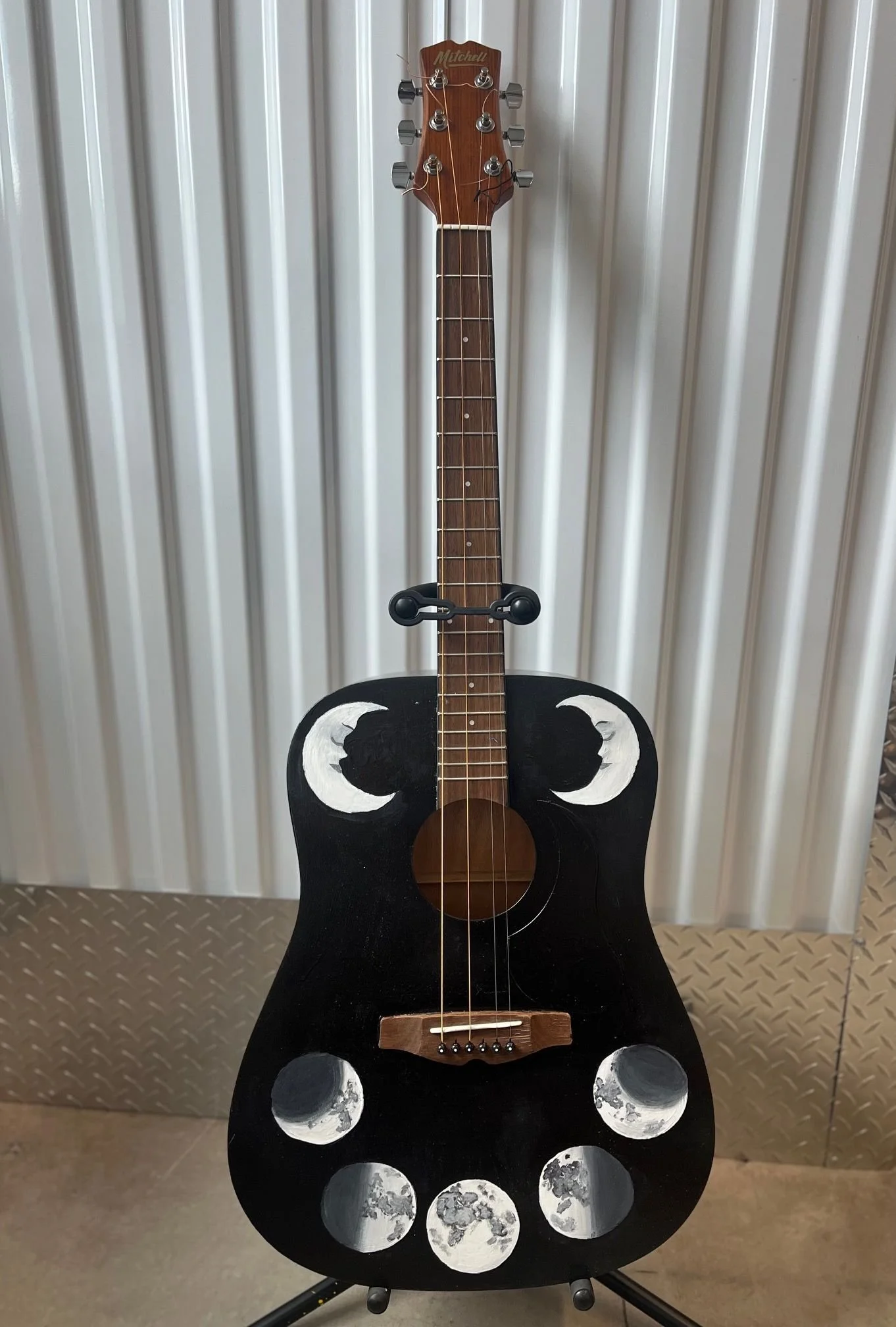 A black acoustic guitar with moon phases painted on the body, standing on a guitar stand against a metal wall.