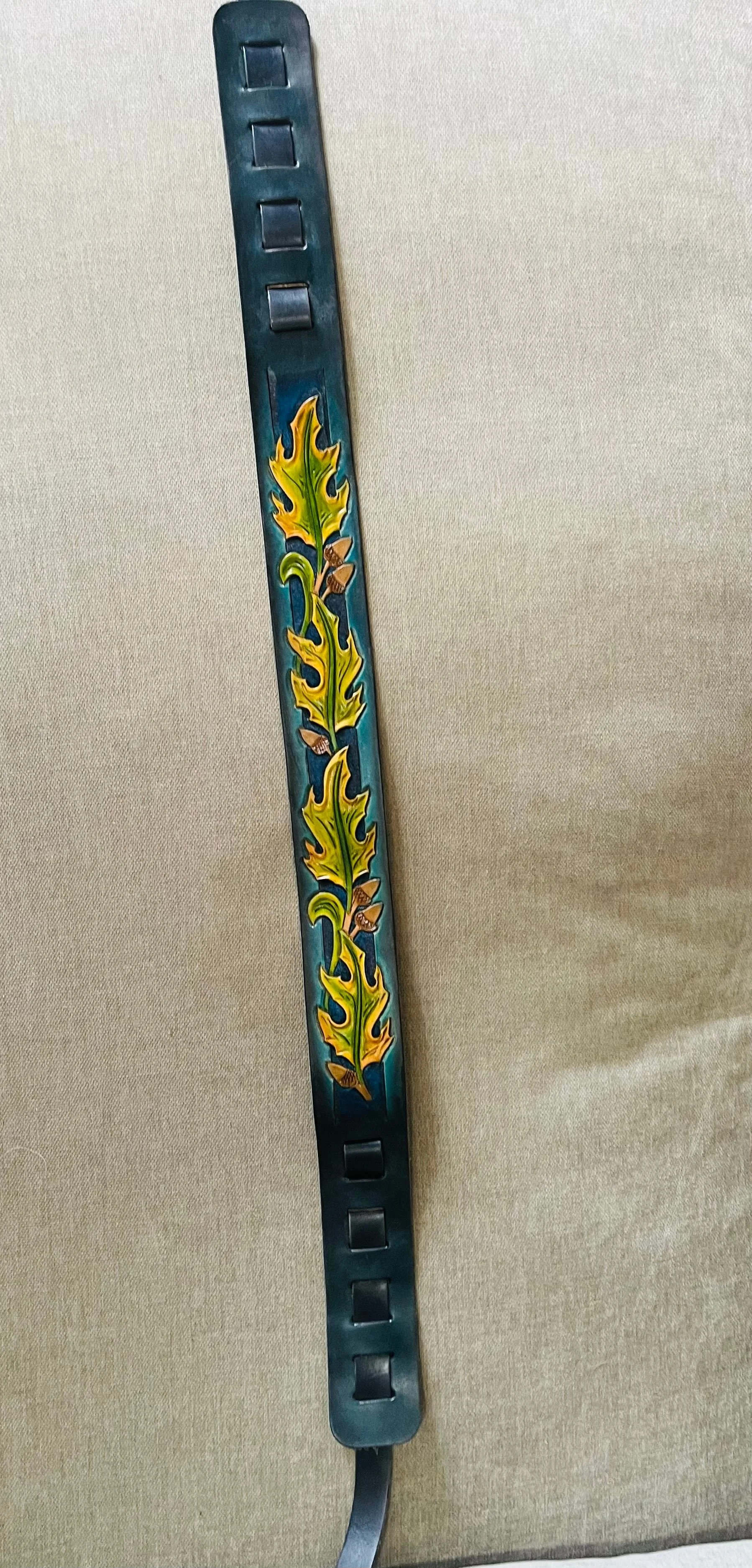 A black metal wristband with a horizontal rectangular design, decorated with colorful oak leaf patterns in yellow and green, with small brown acorns.