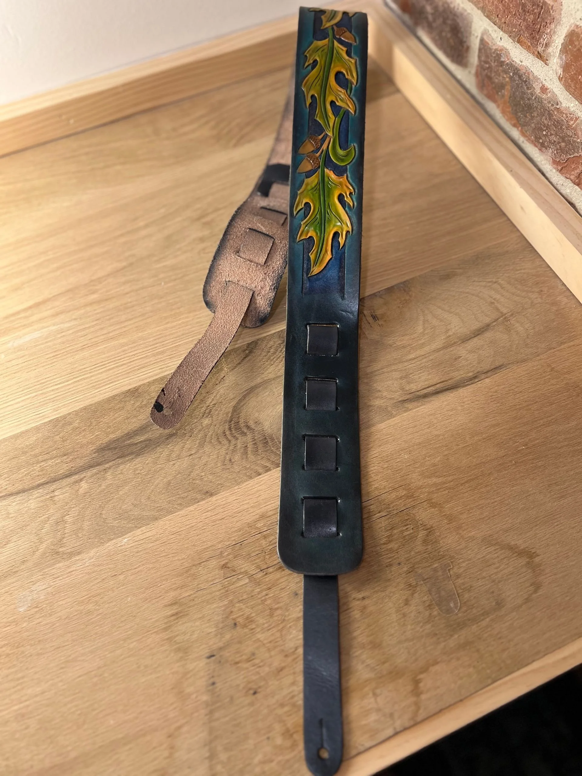 A leather guitar strap with a hand-painted oak leaf and acorn design, resting on a wooden surface with a brick wall in the background.