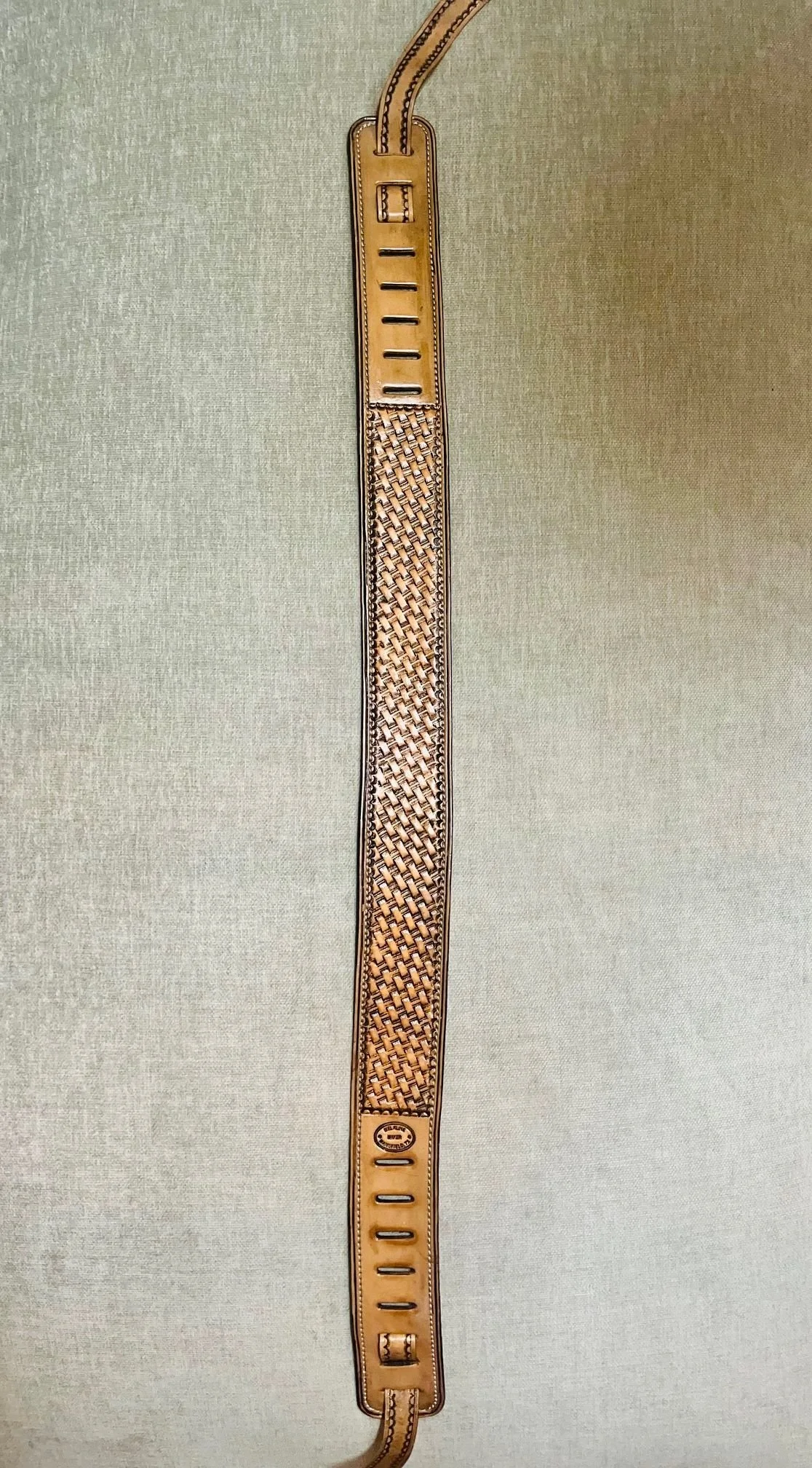 A beige leather belt with a woven pattern in the center, hanging vertically on a light-colored fabric background.