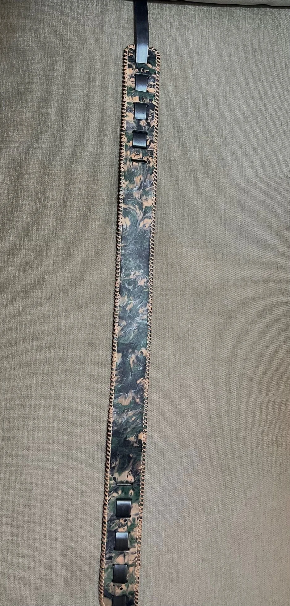 A watch strap with a marbled pattern in earth tones, featuring black links and a braided border, hanging from a black clasp against a beige fabric background.