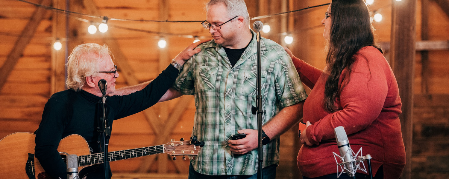 Three people in a cozy wooden room are gathered around a microphone. One person is playing guitar, another is touching the guitarist's shoulder, and the third is standing nearby. There are string lights hanging overhead.