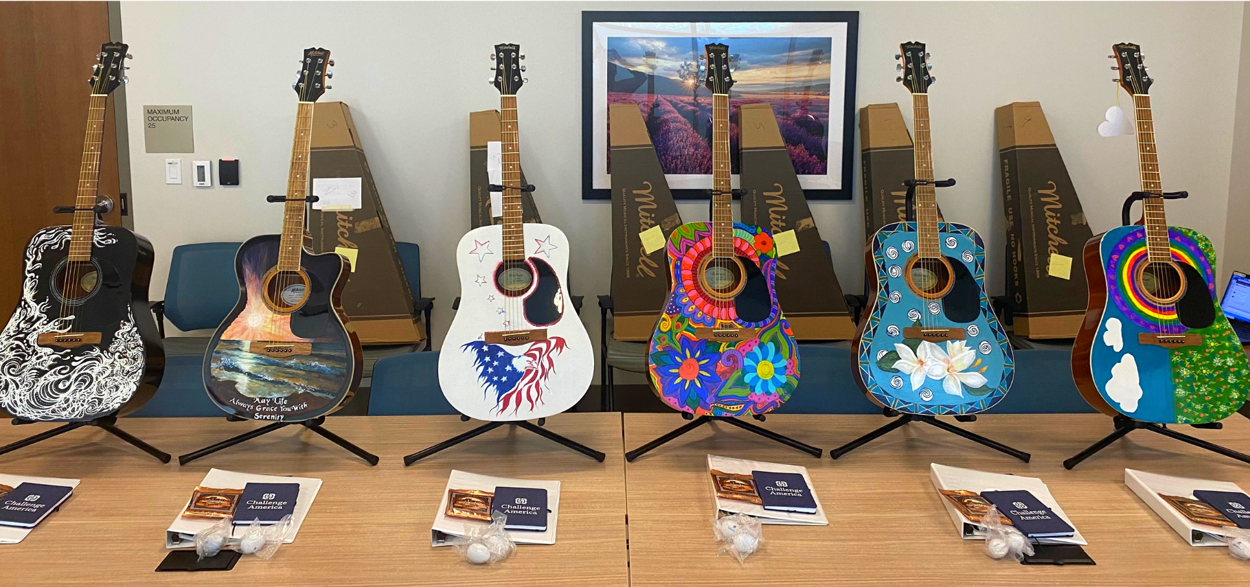 Six decorated acoustic guitars on display, each with unique art designs, placed on stands in front of framed photographs and boxes.