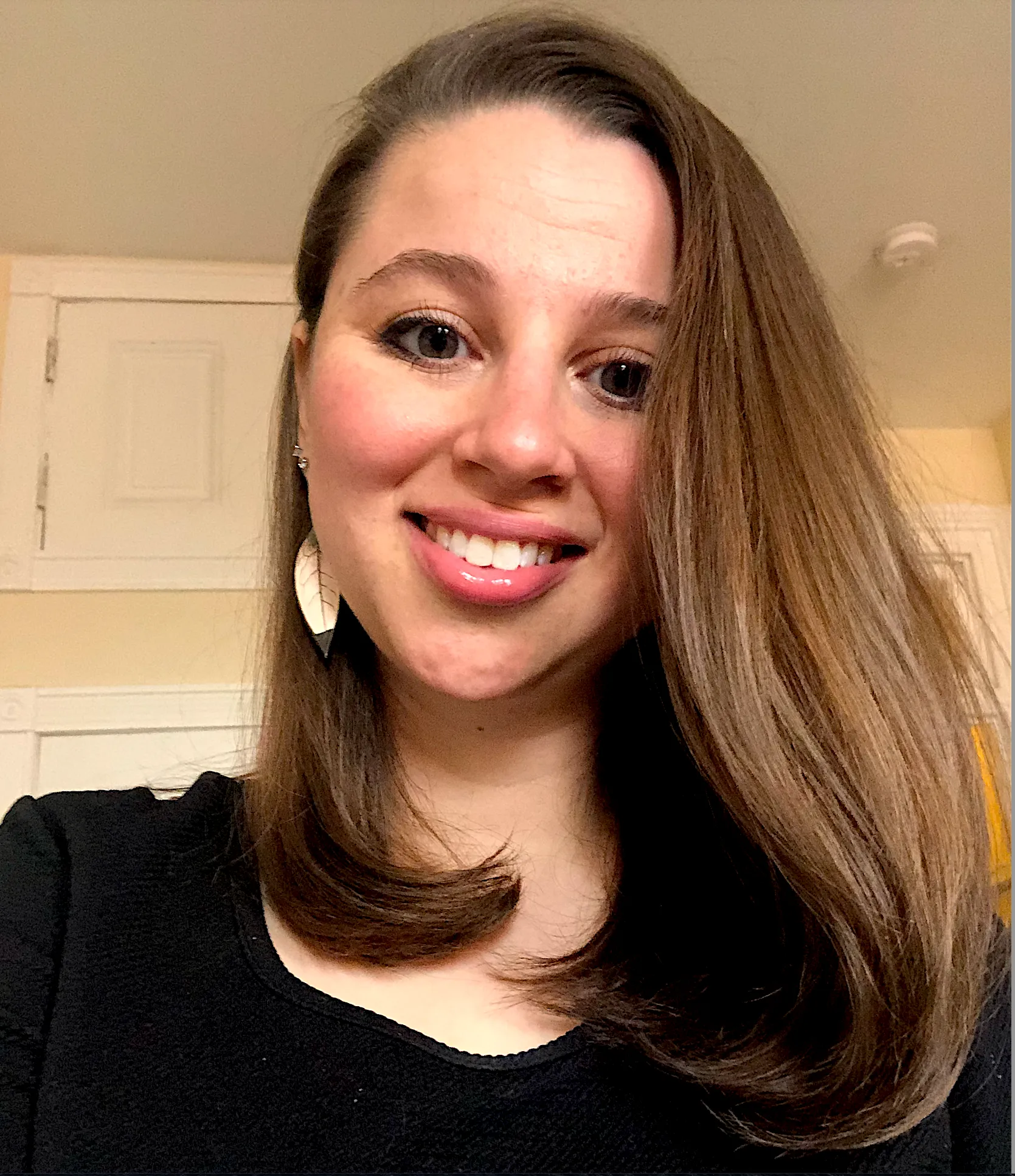 A smiling woman with brown hair, wearing earrings and a black top, in an indoor setting with beige walls and a white door in the background.