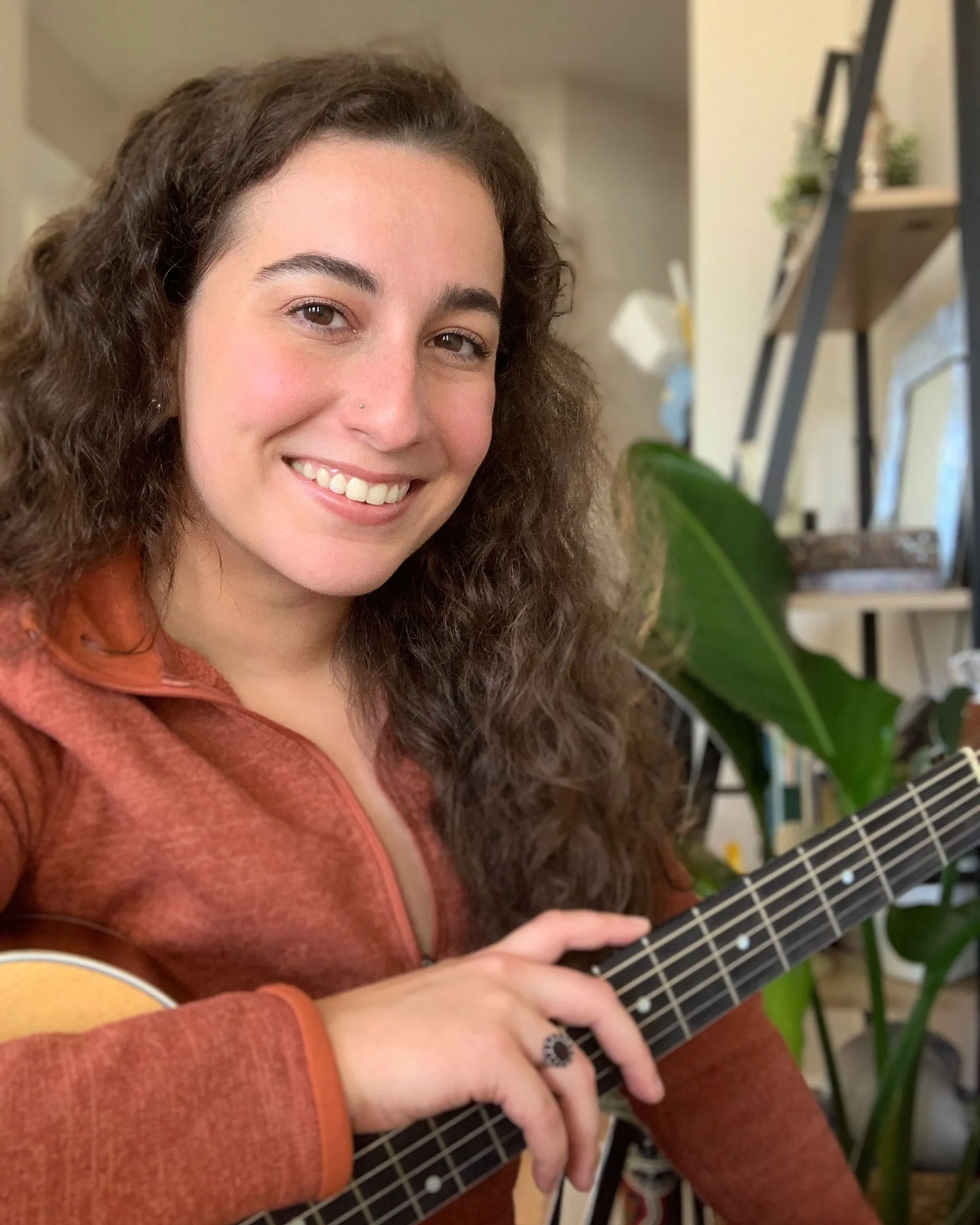 A young woman with curly brown hair smiling and holding a guitar in a cozy indoor space with plants and shelves in the background.