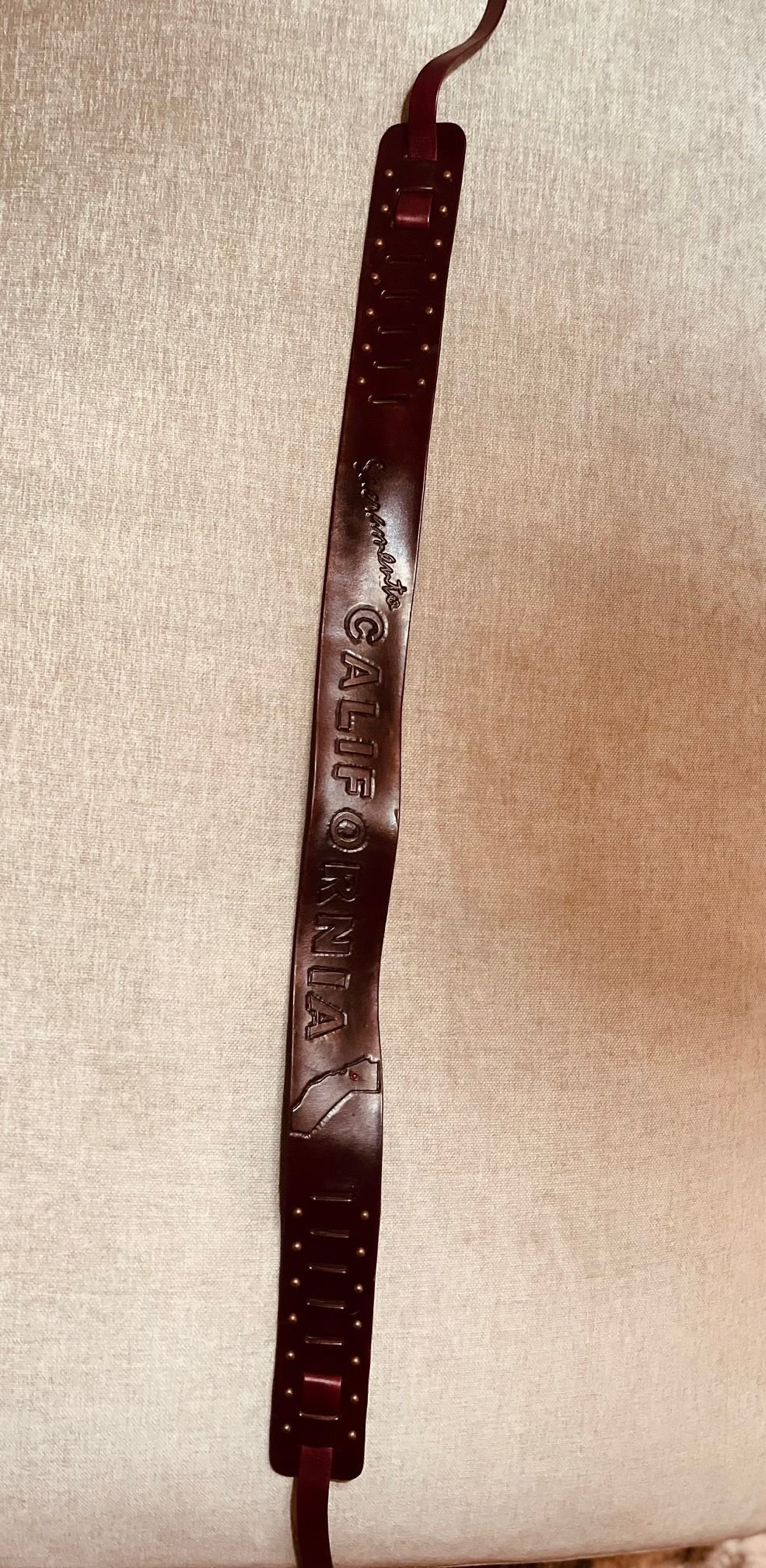 Brown leather wristband with embossed text that reads 'GALLIFORNA', attached to a red strap, hanging on a beige textured wall.