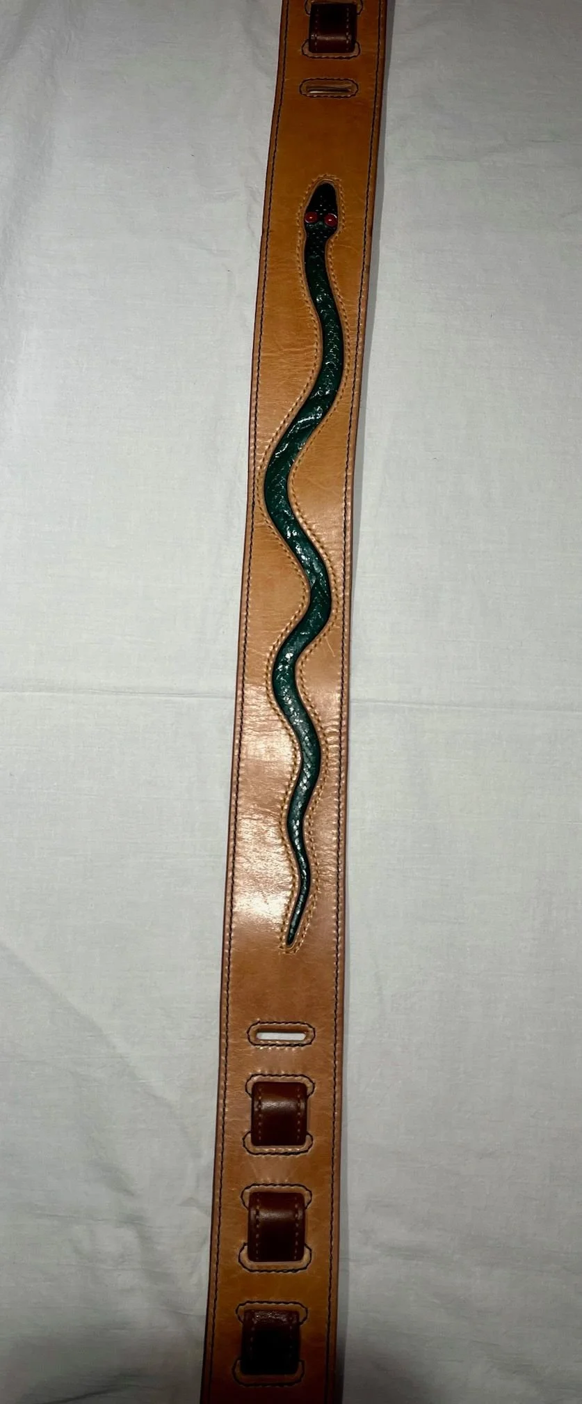 A leather belt with a black snake design on it, featuring red eyes, lying on a light-colored fabric surface.