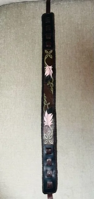 A black leather belt with decorative pink and white flowers and green leaves, laid flat on a beige surface.