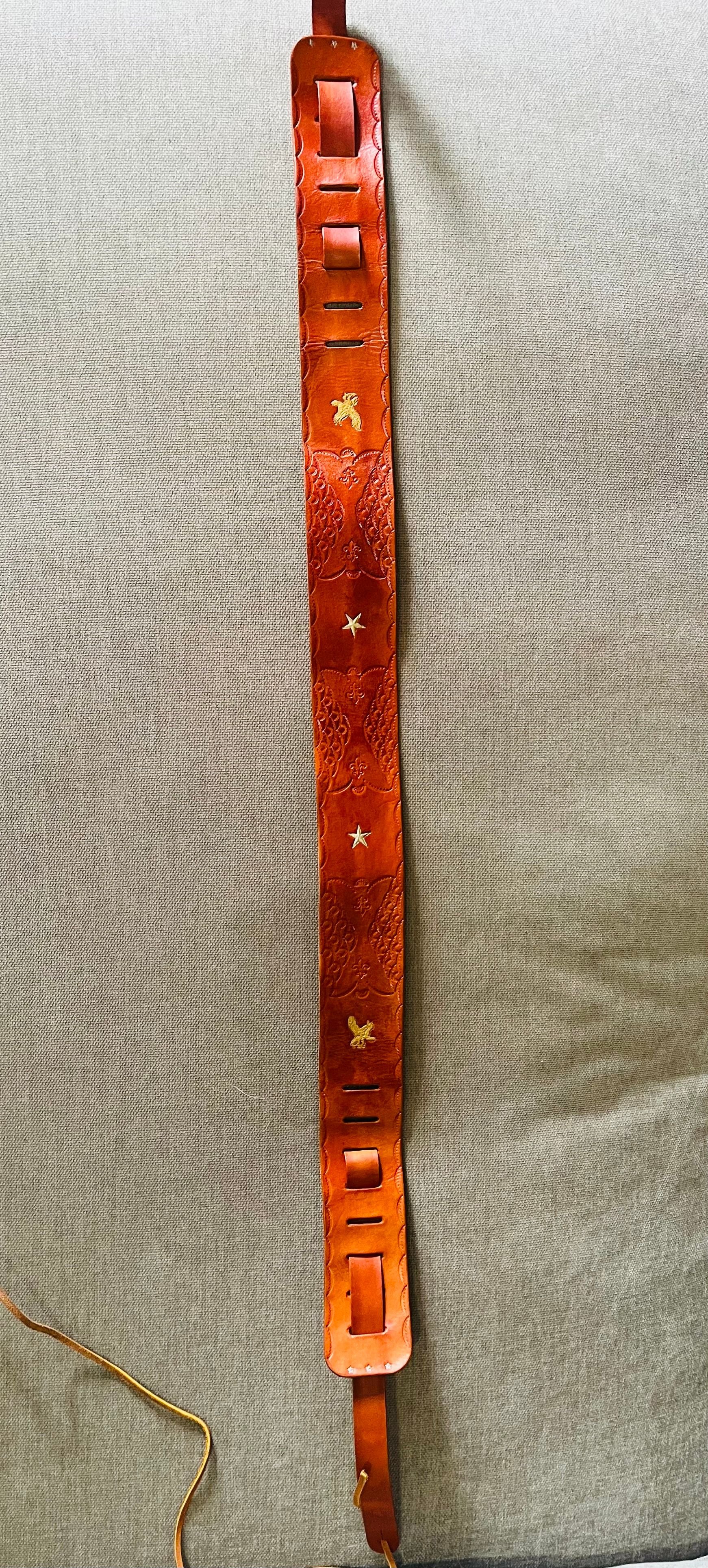 A decorative brown leather belt with engraved floral patterns, gold eagle and star accents, and multiple buckle holes, laid flat on a beige surface.