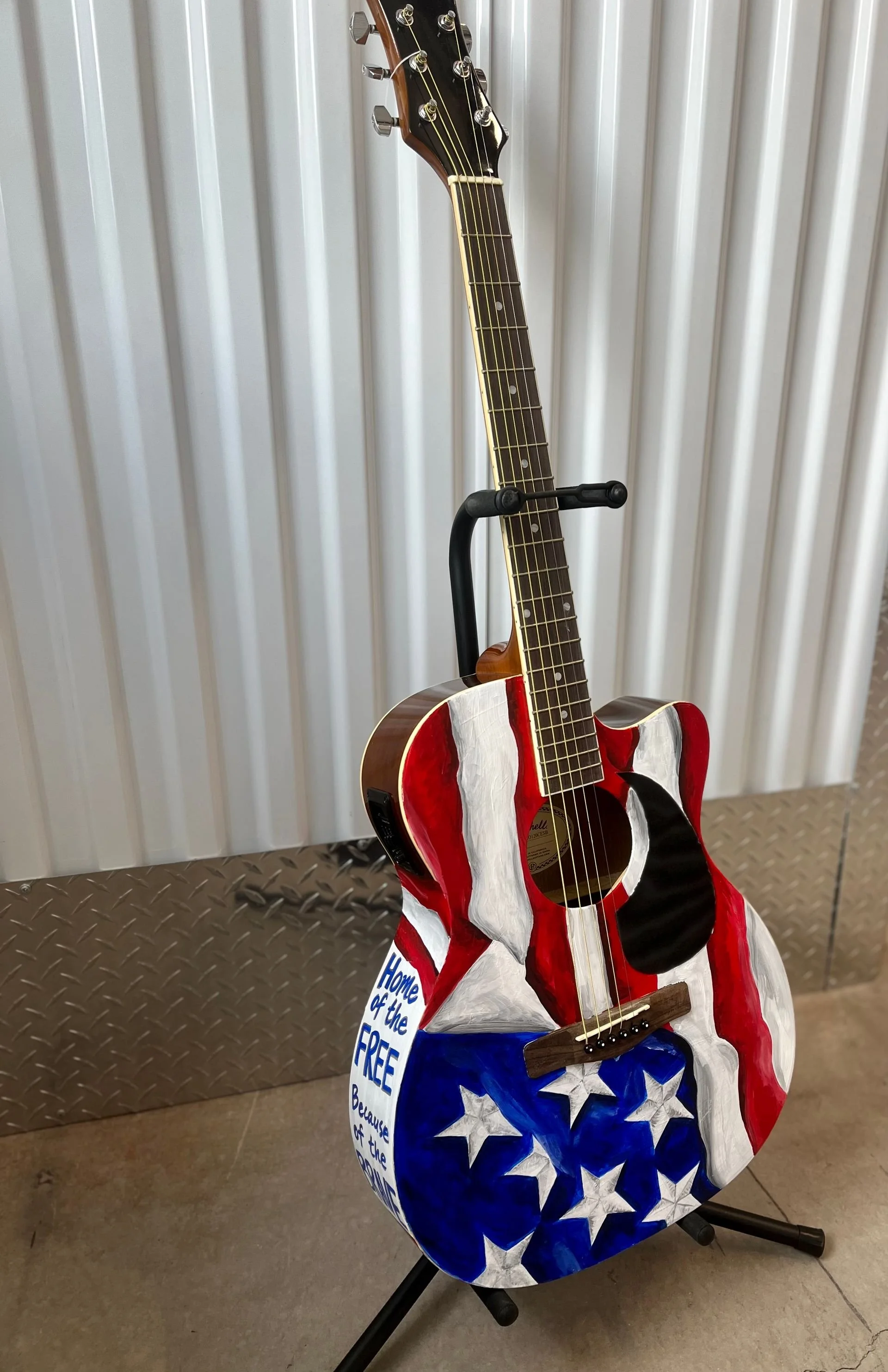 An acoustic guitar with a patriotic design painted on it, featuring red, white, and blue with stars, resting on a black stand.