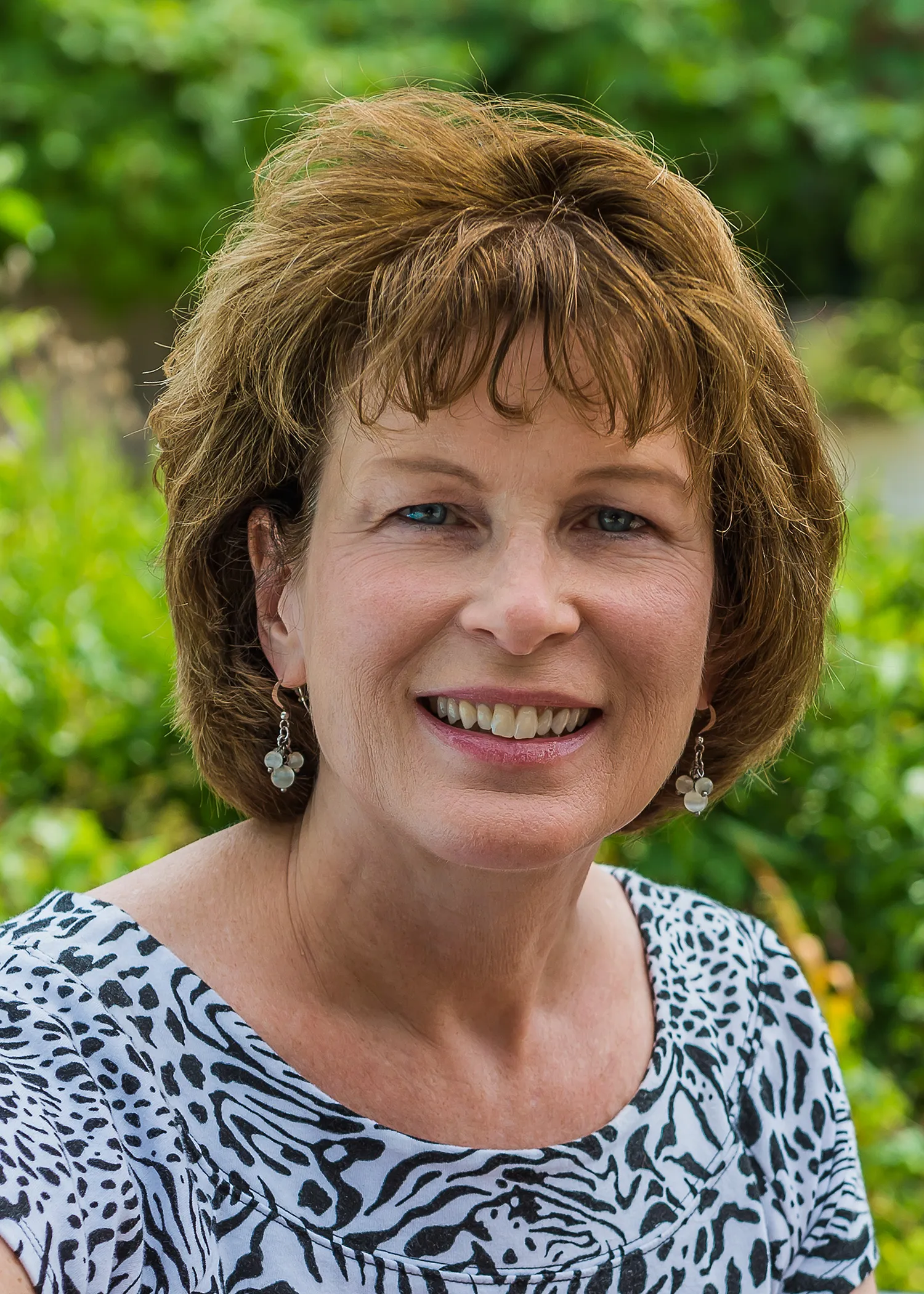 A middle-aged woman with short, brown, wavy hair smiling outdoors, wearing a black and white animal print top and pearl earrings.