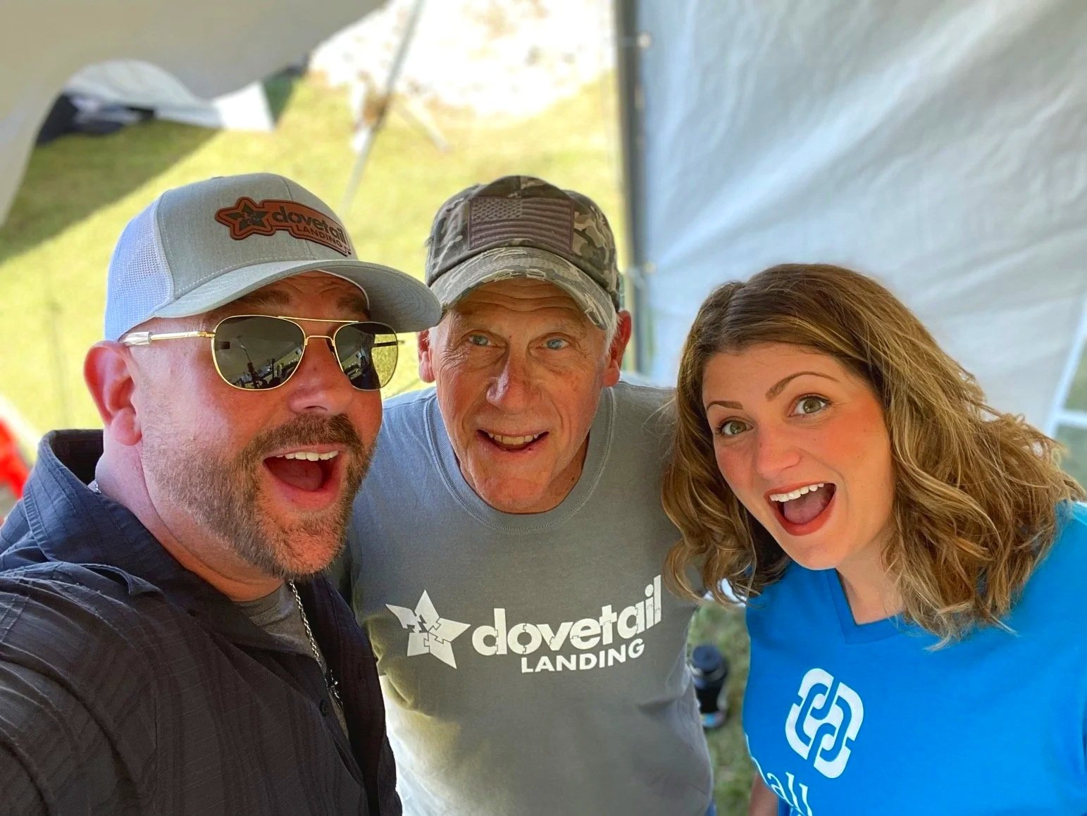 Three people taking a selfie outdoors, smiling and looking happy, with a tent in the background.