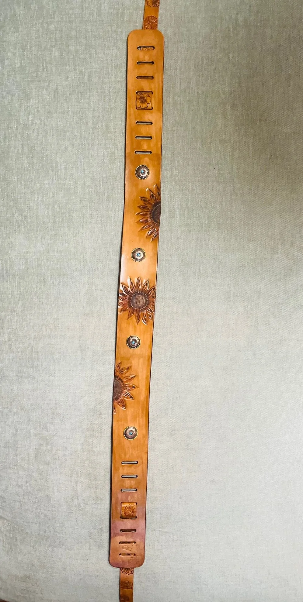 Leather strap with sunflower and floral embossed designs, metal snaps, and slot holes for adjustments, hanging against a textured wall.