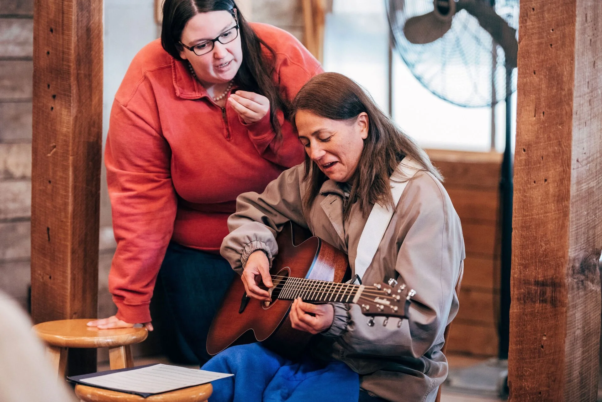 Two women, one sitting and playing a guitar, the other standing and watching, inside a rustic wooden room.